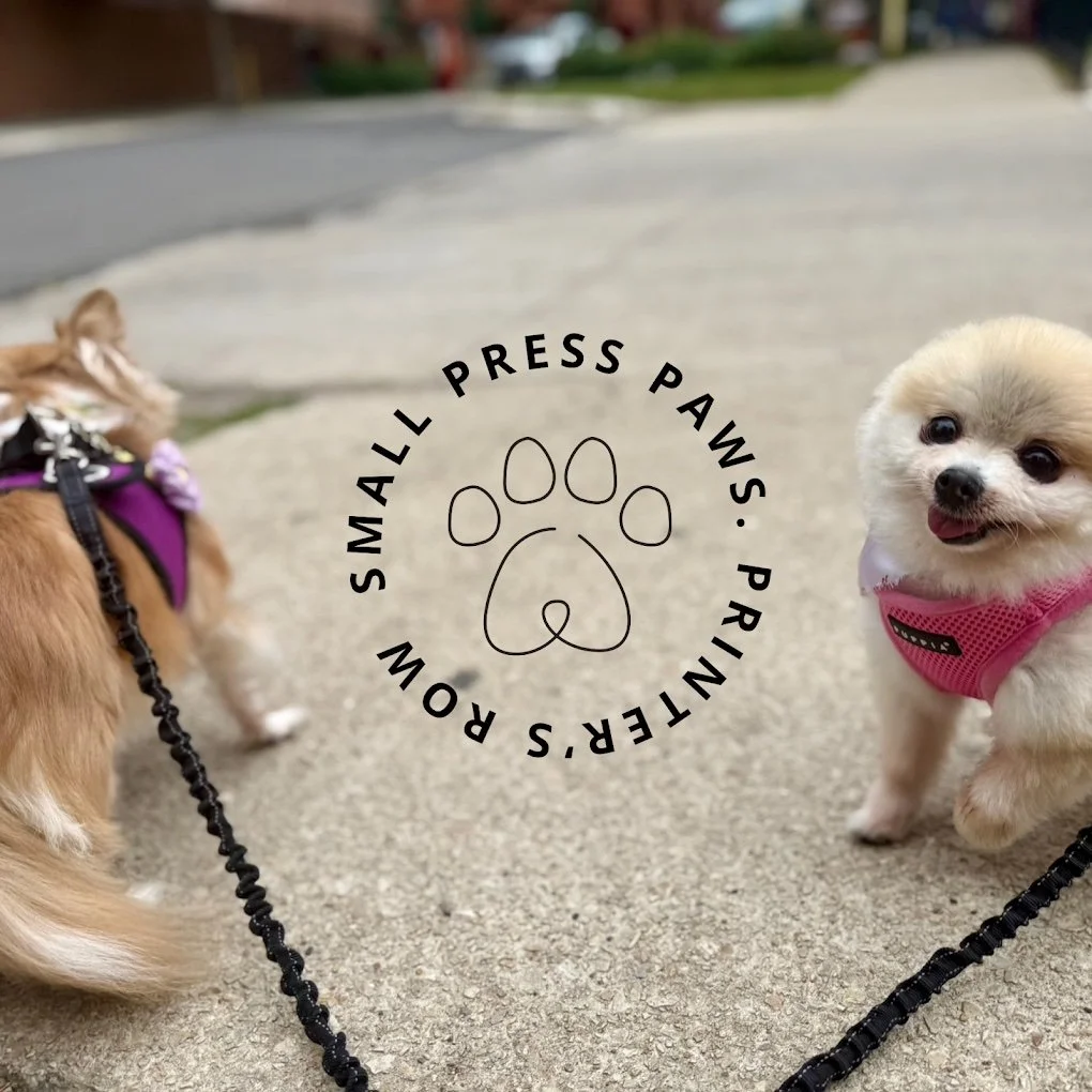 Two small dogs on leashes talking outdoors on a sidewalk, with a logo of a paw print and text reading 'Small Press Paws. Printer's Row' overlaid in the center.