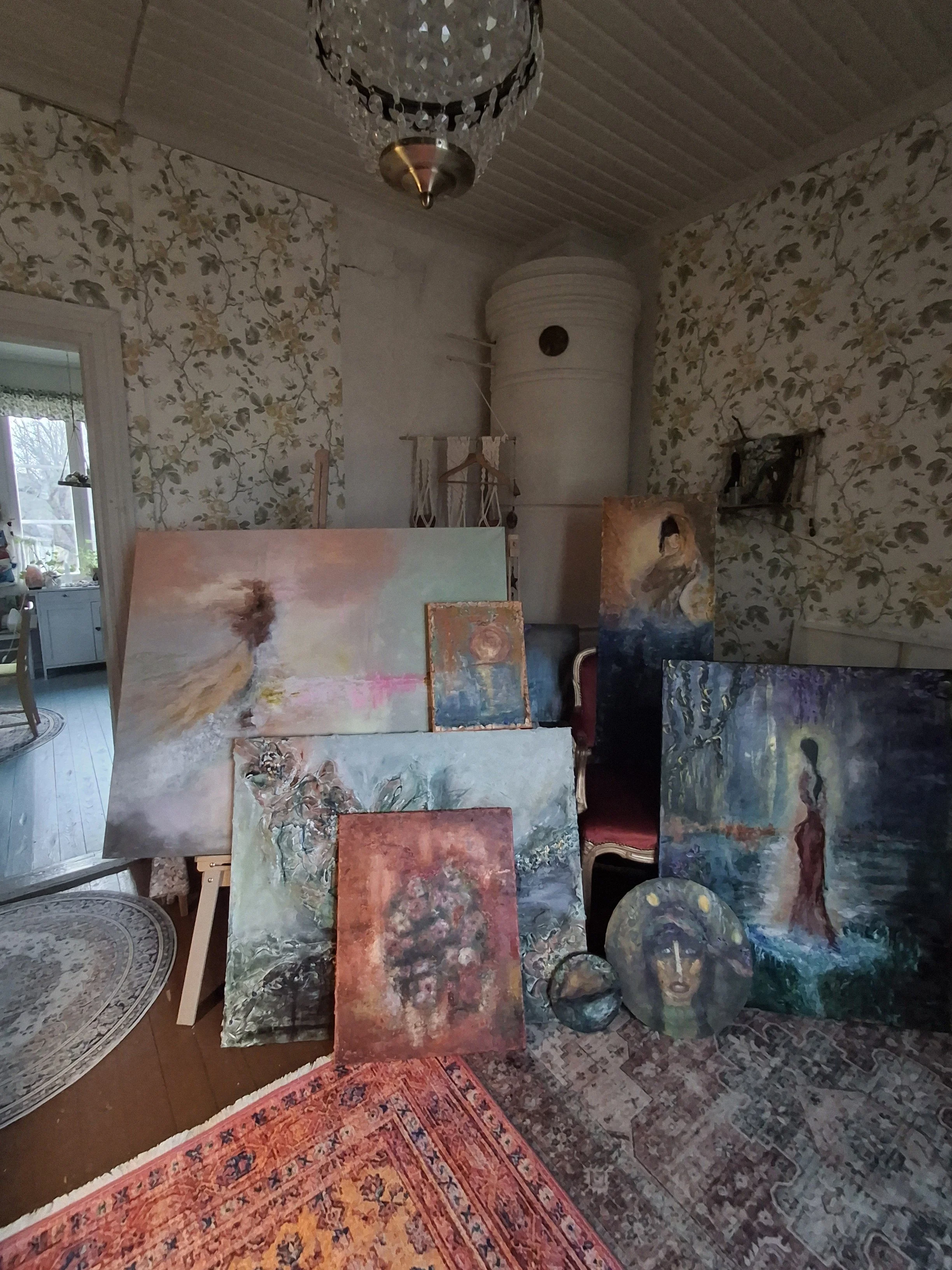 Kuvassa on tauluja ja maalauksia huoneessa, jossa on vanhanaikainen sisustus. Pönttöuuni. Akryylimaalauksia ateljeessa. Ruusutapetti. Old house decor. Vanhan talon ateljee ja sisustus. Beautiful old ateljee. Taidestudio. Artwork. Art studio. Atelier