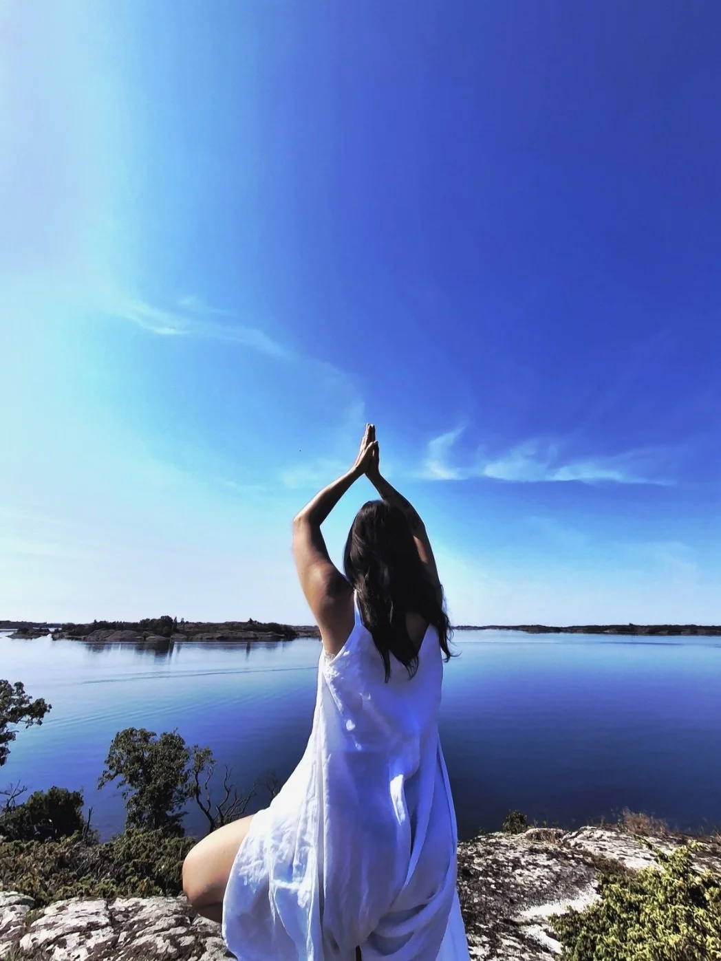 Nainen joogaamassa upeassa maisemassa, korkean kallion reunalla meren äärellä, taivas aivan kuin jakautuu meditatiivisessa asennossa olevista  käsistä valkoiseen pilveen.
