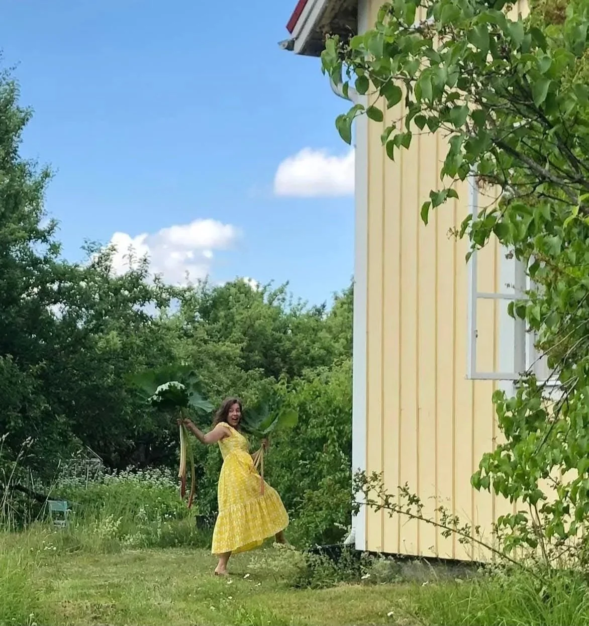 Vihreän pihan keskellä nainen hymyilee ja tanssii kesällä, hänellä on keltainen kukkamekko ja suuri raparperin lehti kädessä. Vanhan talon puutarha. Ateljeen kaunis vanha piha. Atelier AnnaMaya. Old Villa and ateljee. Artist in her old garden.