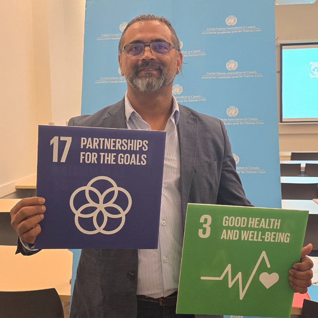 A man in a suit holding signs referring to sustainable development goals: one sign is purple with white text that reads '17 Partnerships for the Goals' with a logo, and the other sign is green with white text that reads '3 Good Health and Well-being' with a health icon. The background features a blue UN banner.