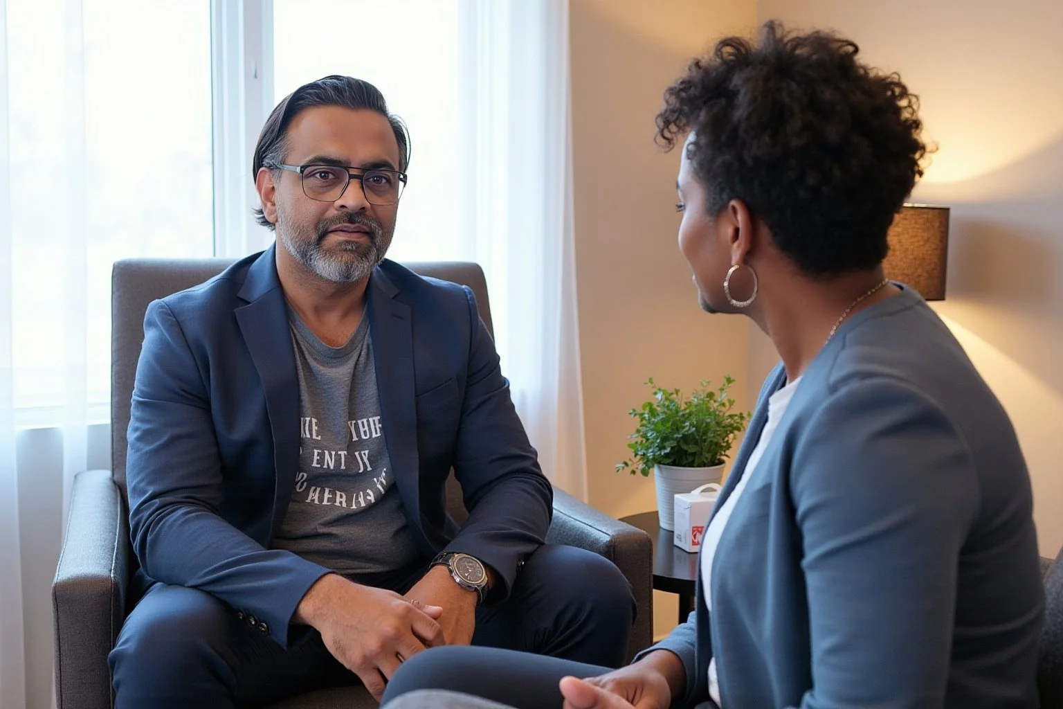 A man and woman having a serious conversation in a well-lit room. The man wears glasses, a blue blazer, and a gray T-shirt, sitting on a gray armchair. The woman, with short curly hair and hoop earrings, wears a gray blazer, sitting across from him. There is a plant on a table between them, and a lamp on the wall.
