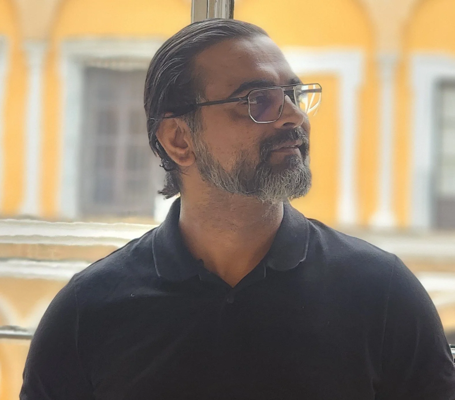 Side profile of a man with salt and pepper hair, beard, and glasses, wearing a black collared shirt, sitting outdoors with a yellow building in the background.