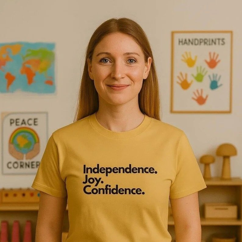 Young woman with long red hair smiling in classroom with educational posters about handprints, peace, and world map in the background, wearing a yellow t-shirt with the words "Independence. Joy. Confidence."