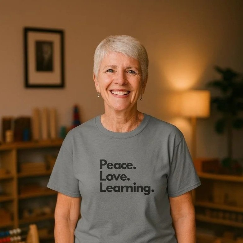 An older woman with short white hair, smiling, wearing a gray T-shirt that says 'Peace. Love. Learning.' standing in a cozy, well-lit room with bookshelves and a lamp in the background.