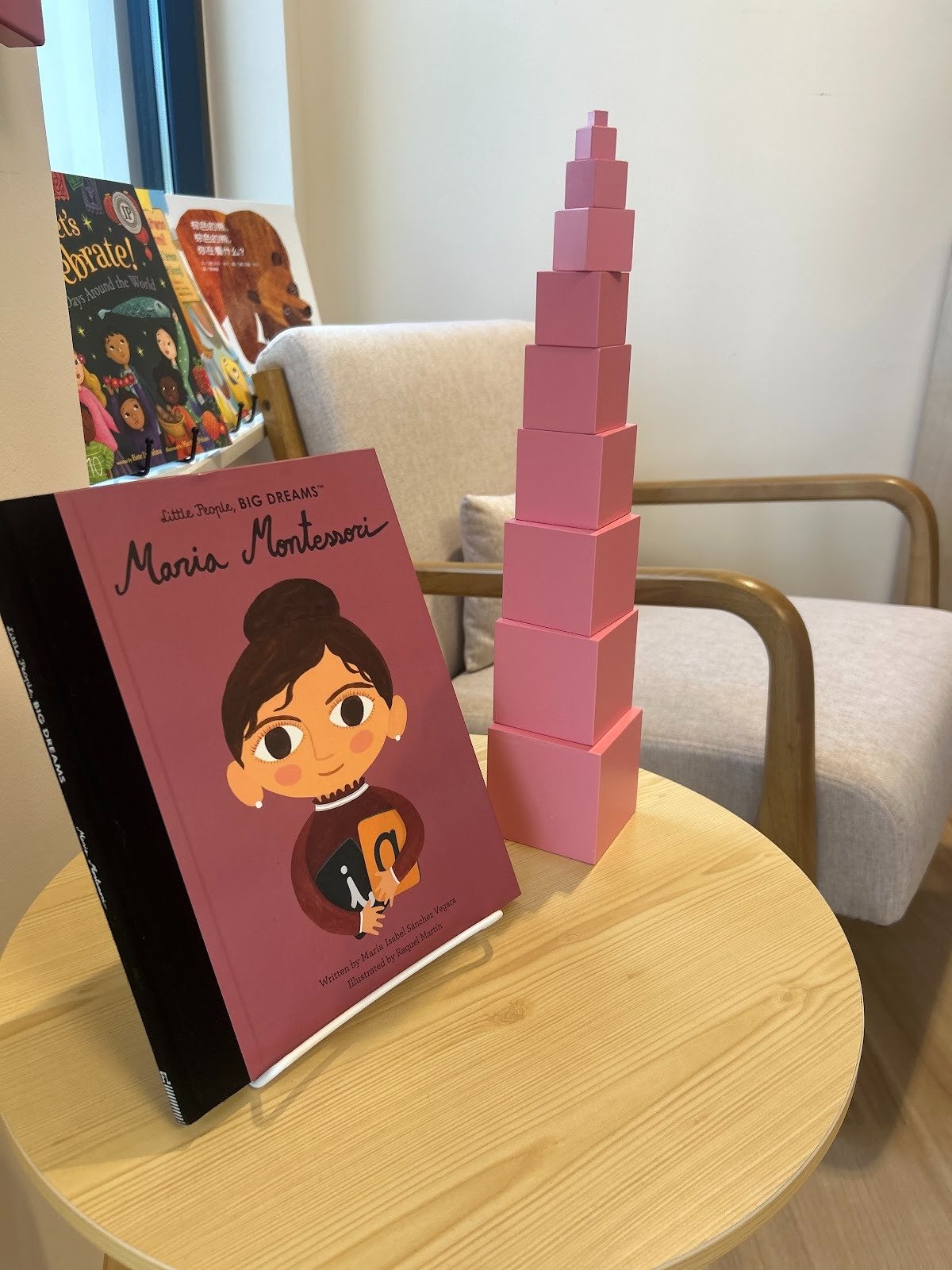 A pink book titled 'Maria Montessori' from Little People, Big Dreams collection displayed on a round wooden table, next to a pink block tower with stacked cuboids getting smaller at the top, and a beige armchair in the background.