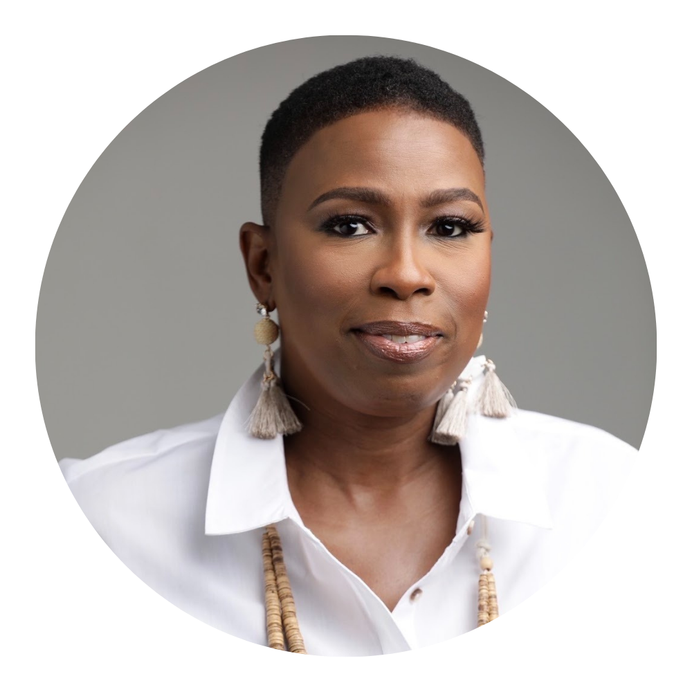 A woman with short natural hair, wearing a white blouse, beige earrings, and layered necklaces, posed against a neutral gray background.
