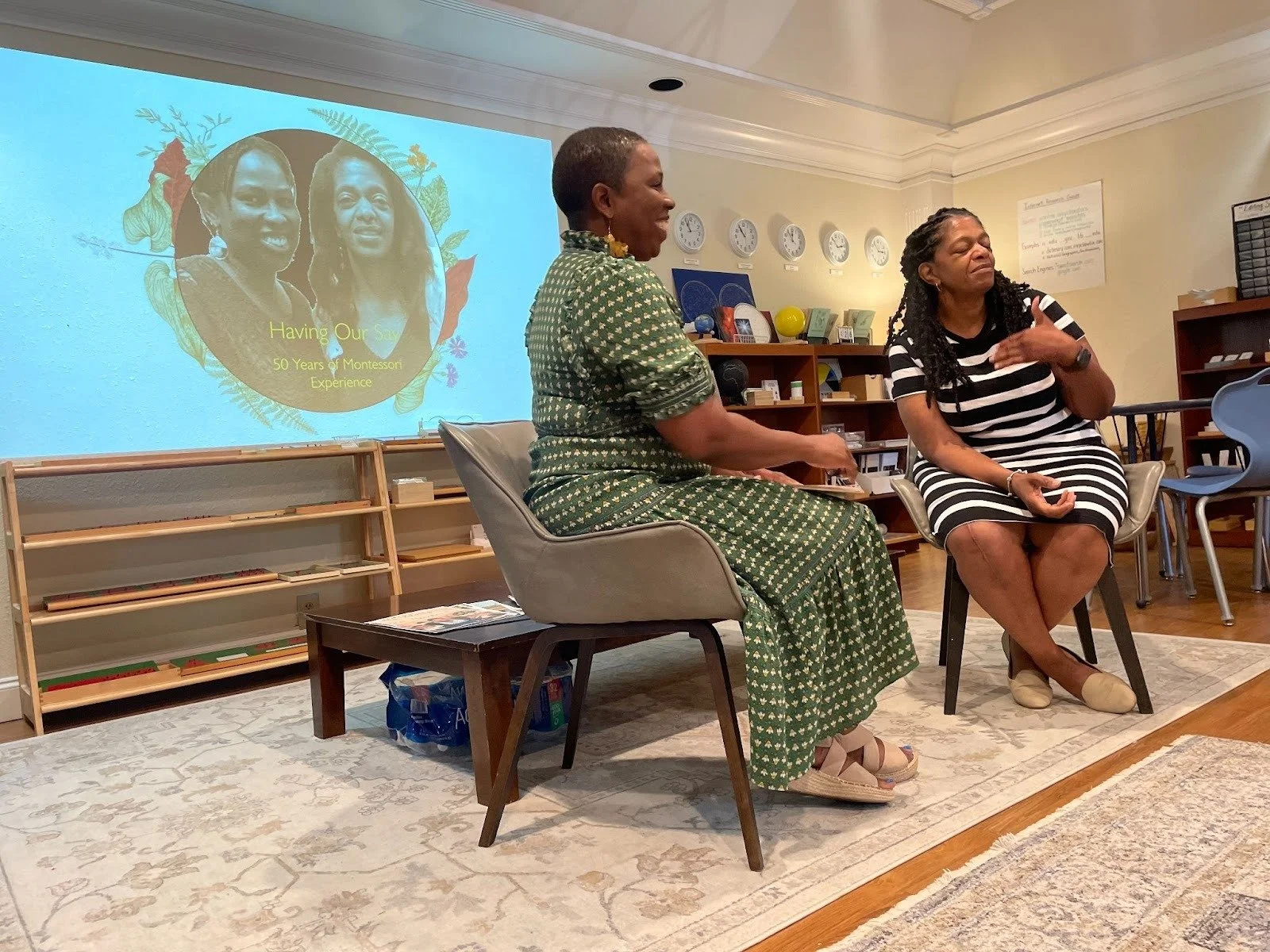 Two women are sitting and talking in a room, with a presentation slide projected on the wall behind them. The slide says, "Having Our Say: 50 Years of Montessori Experience," and features a photo of two women. The room has bookshelves, clocks, and educational materials.