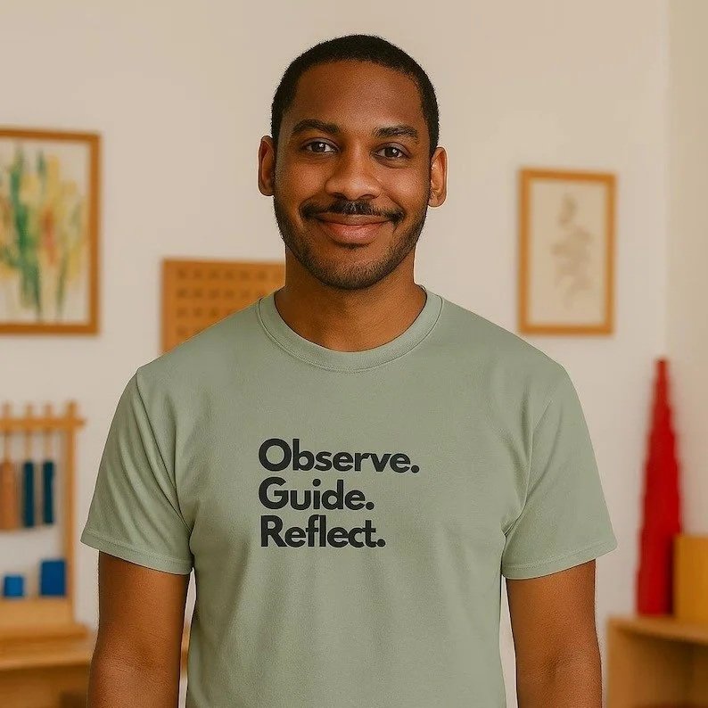 A smiling man standing indoors in a room with framed artwork on the walls, wearing a light green t-shirt with the words 'Observe. Guide. Reflect.' printed on it.