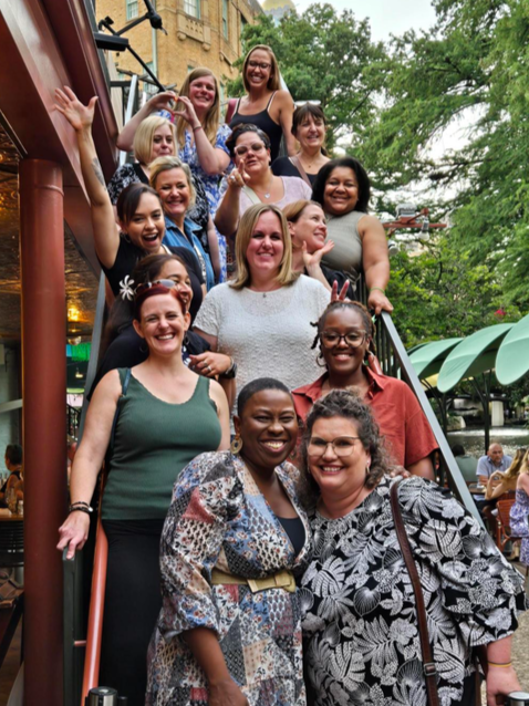 Group of women smiling and posing on outdoor staircase, some making peace signs, with trees and outdoor seating area in background.