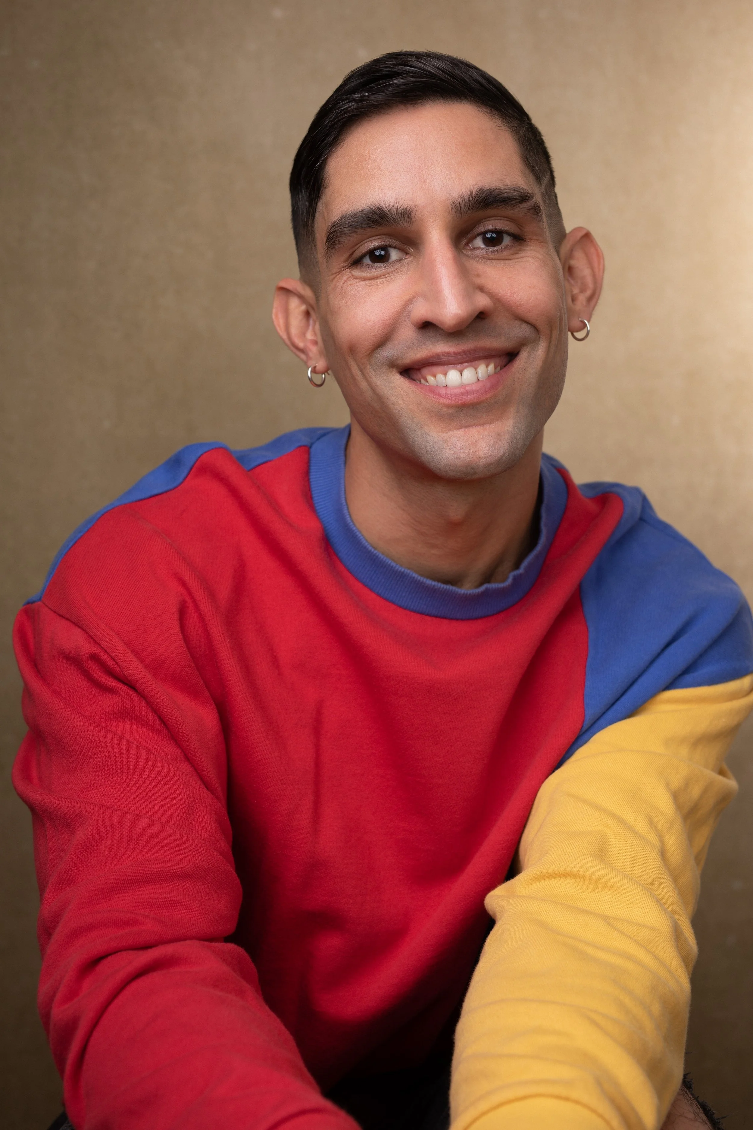 Young man with short dark hair, earrings, and a bright smile, wearing a multicolored sweatshirt in red, blue, and yellow, against a neutral background.