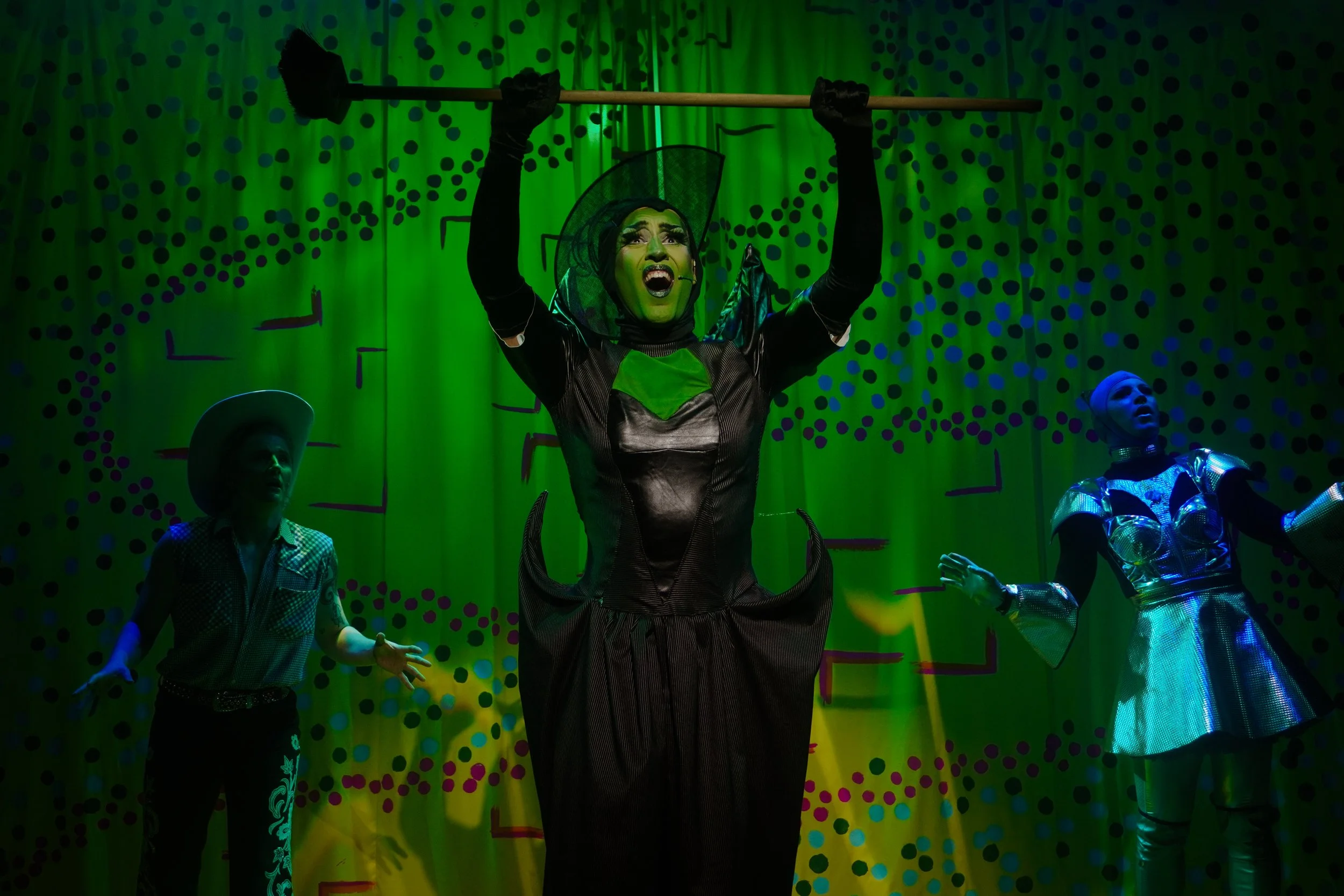 A theatrical performance with actors in colorful costumes, with one central actor dressed as a spider holding a staff above her head, surrounded by two other performers on a vibrant, dotted backdrop.