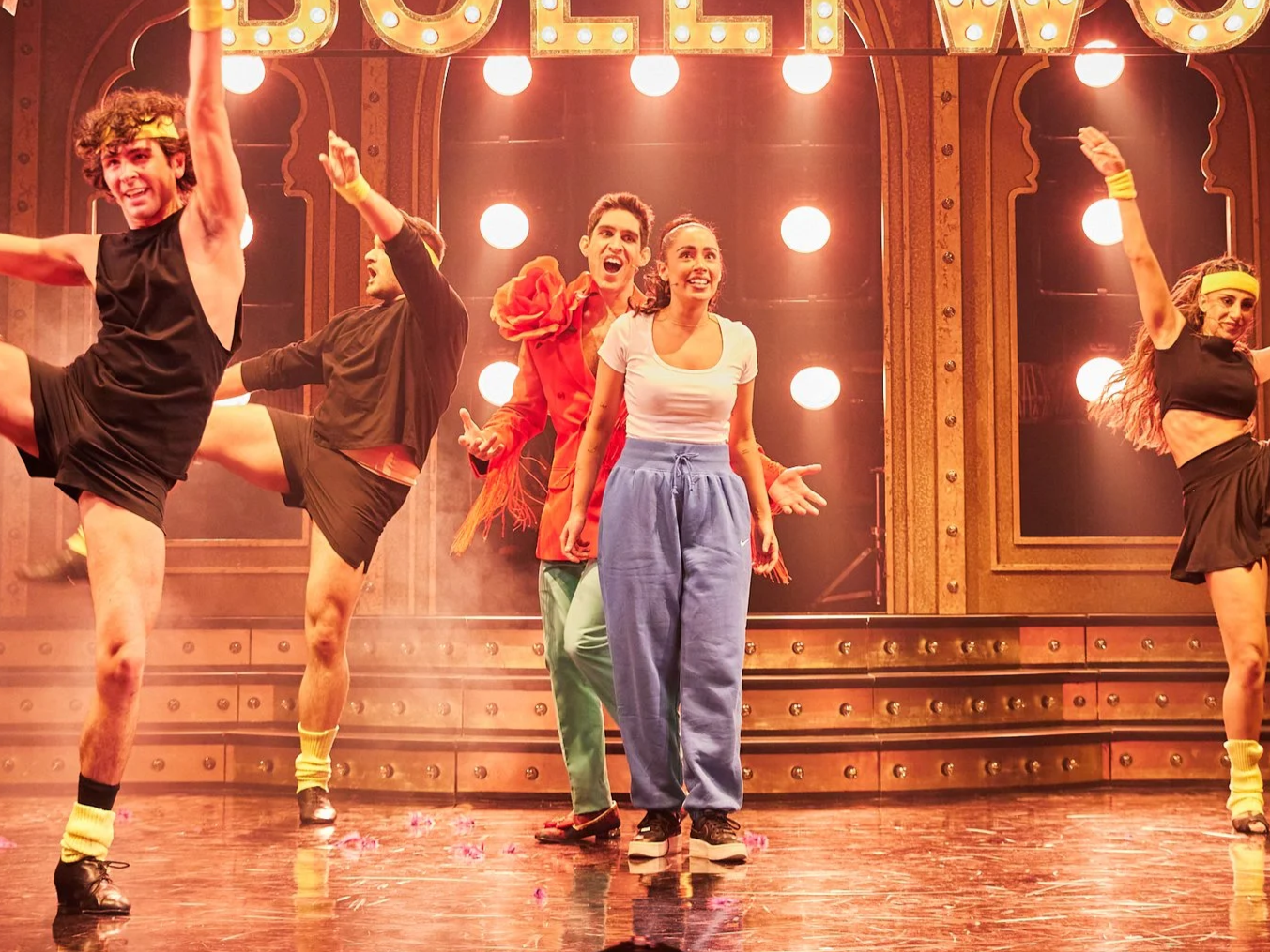 Actors performing on stage with bright lights and a vintage style backdrop.