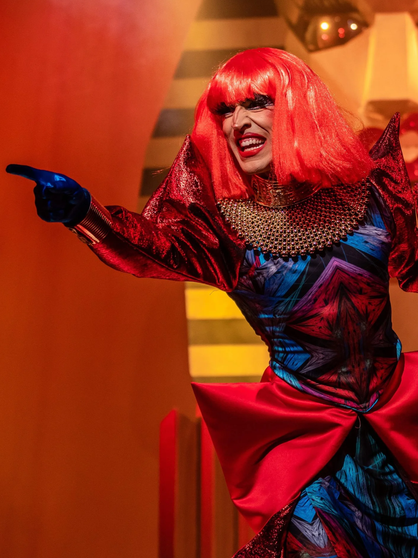 Performer with long red hair wearing elaborate costume pointing and smiling.