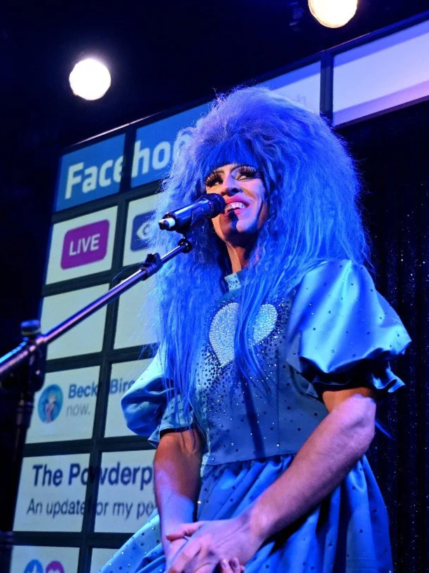 A drag queen with large, blue curly hair performing on stage in front of a backdrop displaying Facebook logos, using a microphone. The performer is dressed in a shiny, blue outfit with a heart design, and is singing or speaking into the mic.