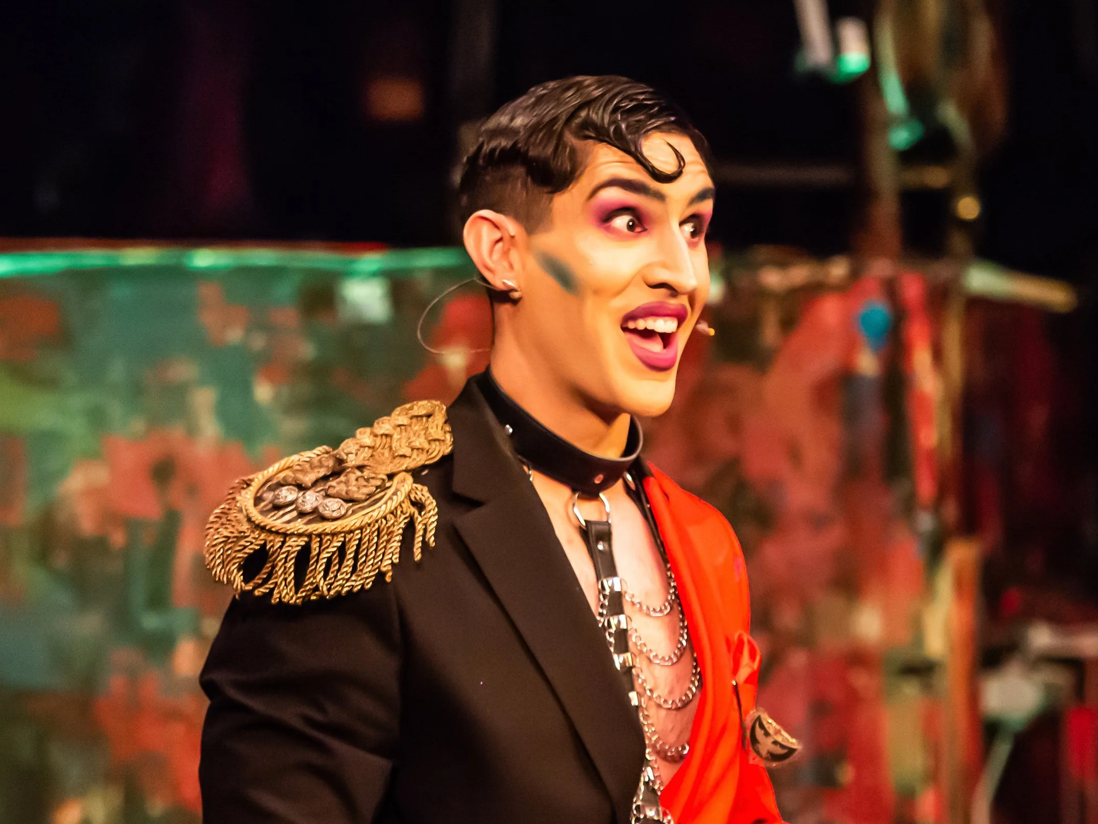 Person dressed in a flamboyant outfit, with a decorated shoulder epaulette, bright makeup, and short, styled hair, performing on stage with colorful lighting and background.