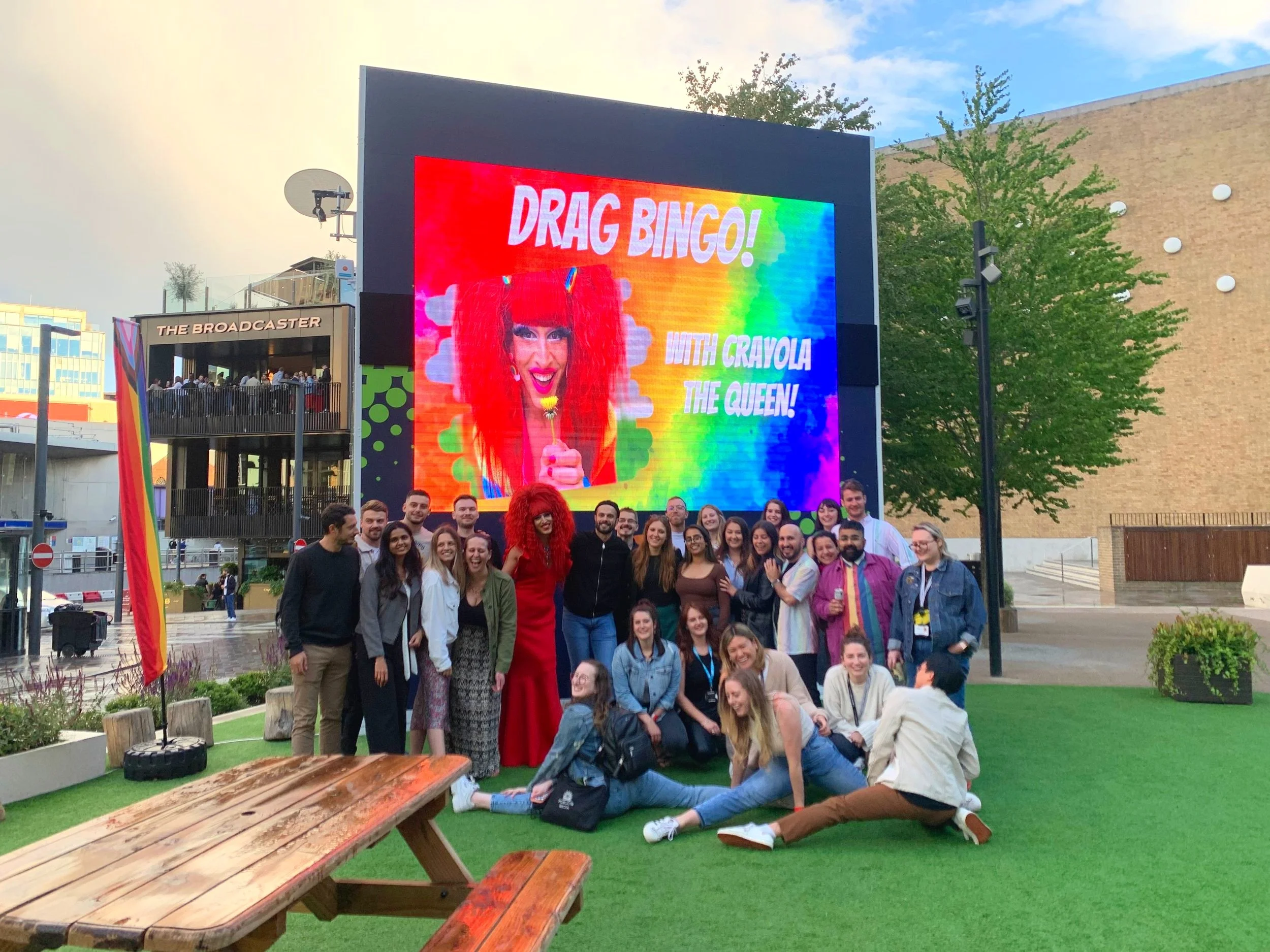 Group of people posing in front of a large screen advertising 'Drag Bingo! with Crayola the Queen!' in an outdoor setting with a pub and seating area visible.