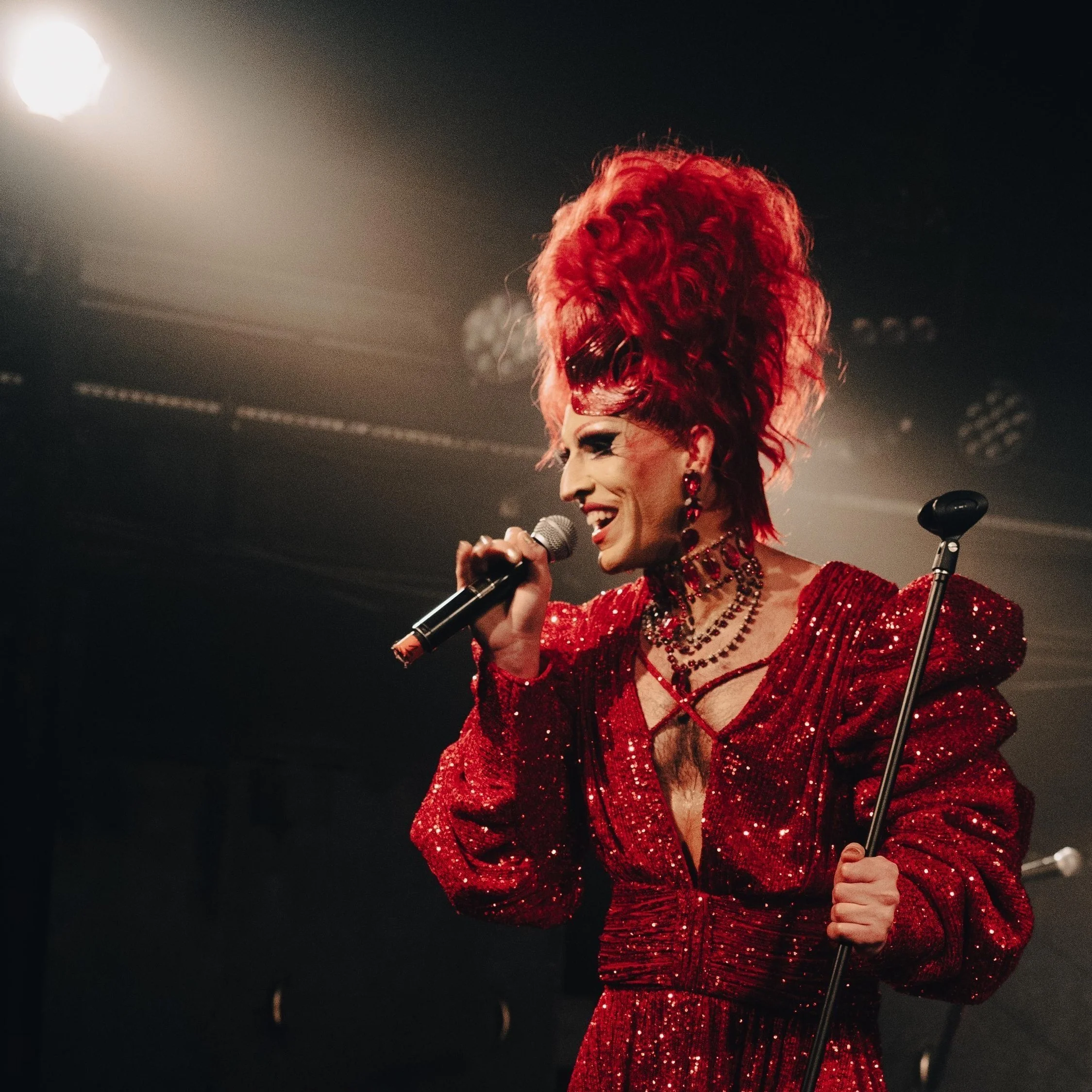 Person dressed as a devil in a red, sequined gown, with red horns and makeup, speaking into a microphone at a pride event decorated with rainbow flags.