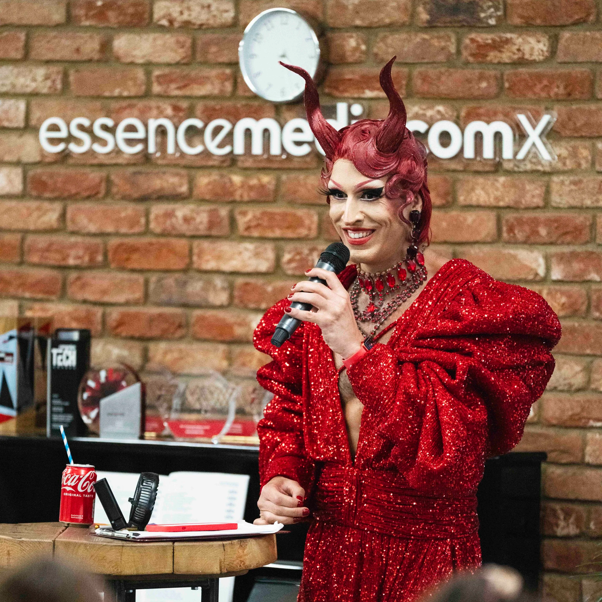 A person dressed as a demon with red hair, horns, and dramatic makeup, holding a microphone and smiling. They are wearing a sparkly red dress and red jewelry, standing in front of a brick wall with a sign that reads 'essencemakeup'.