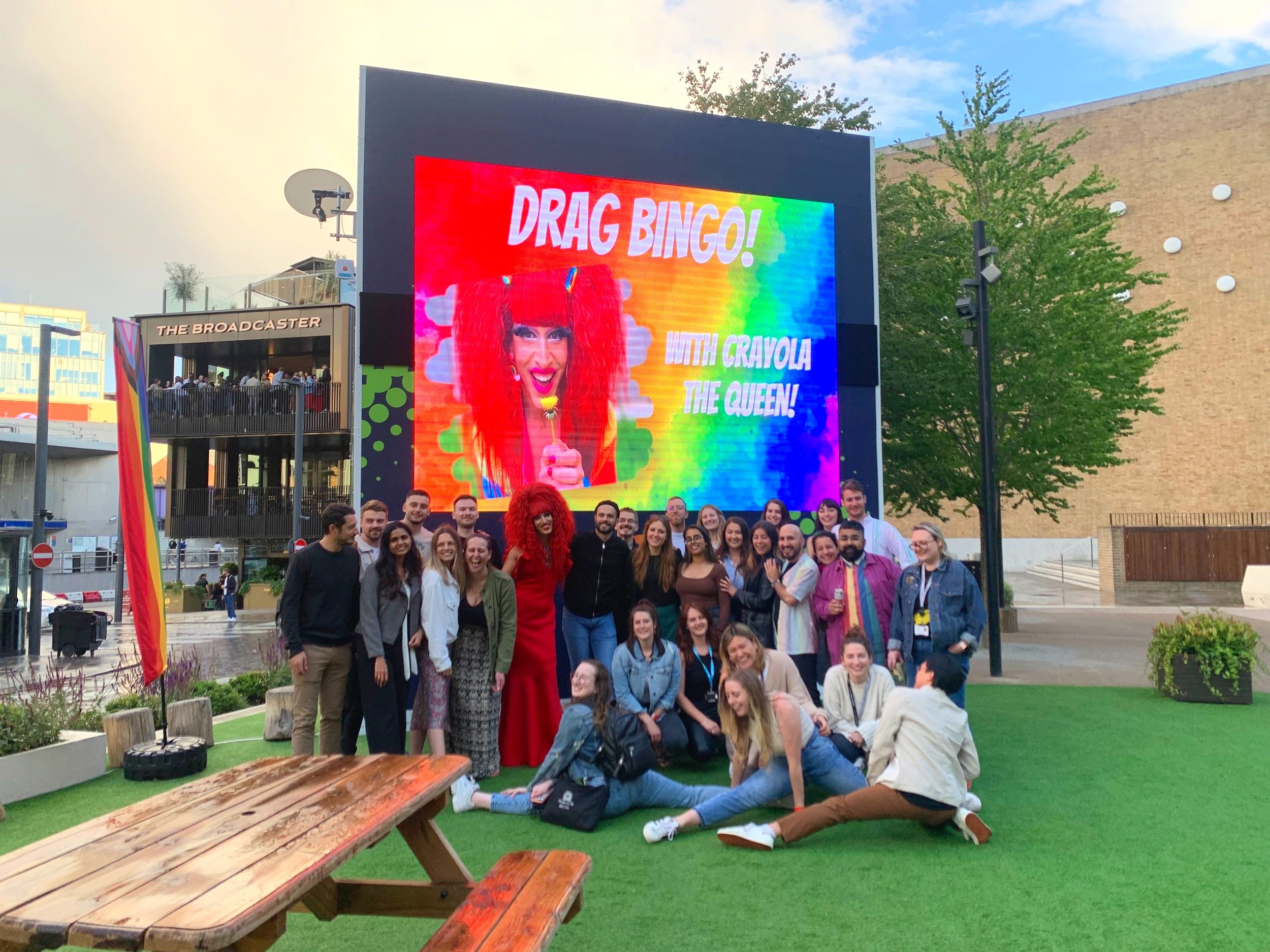 A group of people standing and sitting in front of a large rainbow-colored digital screen outdoors. The screen displays a woman with red hair and a colorful background, promoting an event called Drag Bingo with Crayola The Queen. There are trees, a brick building, and a sign reading 'The Broadcaster' in the background.