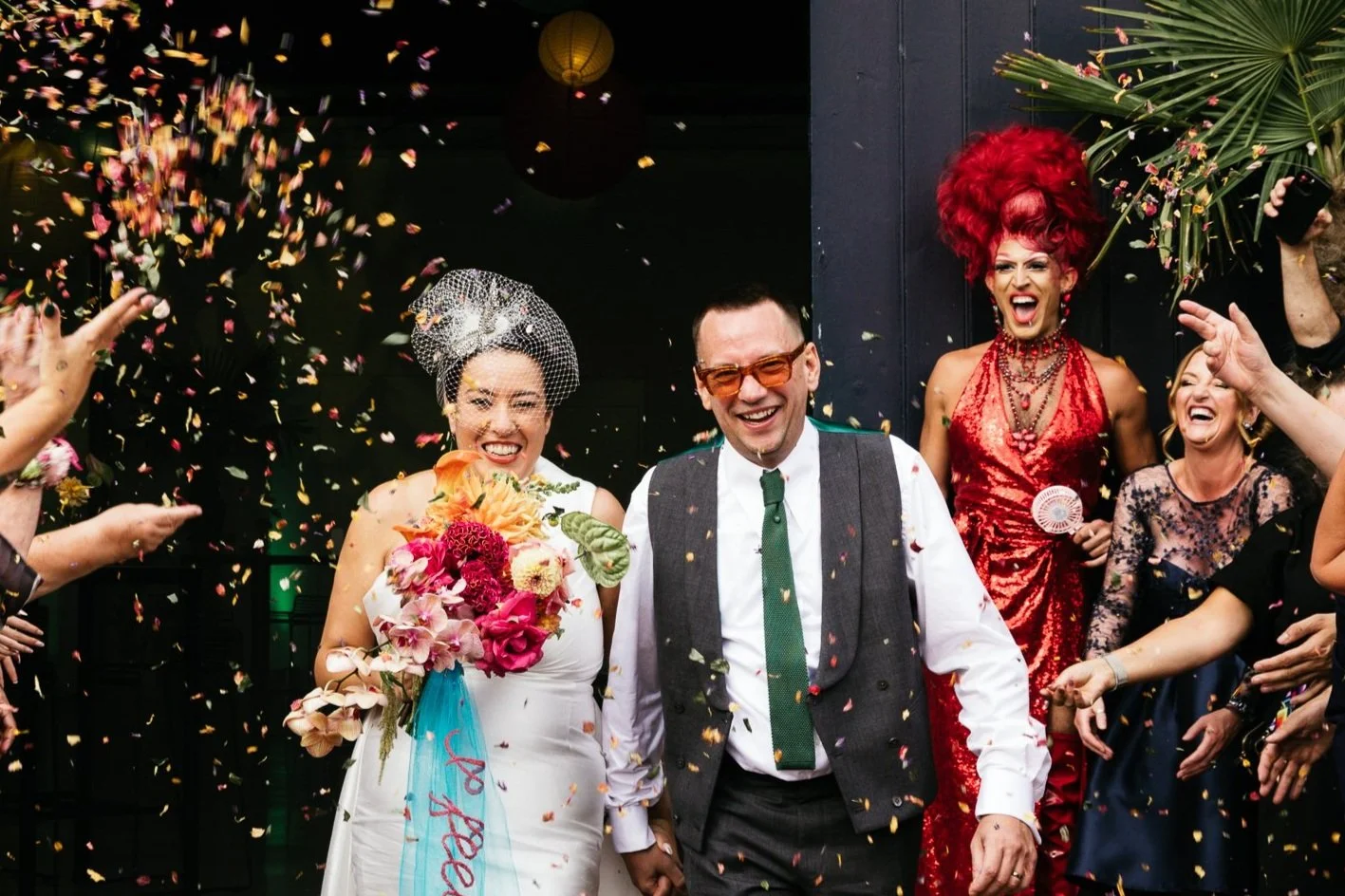 A couple joyfully exiting a venue as guests throw confetti, surrounded by vibrant colors and festive attire.