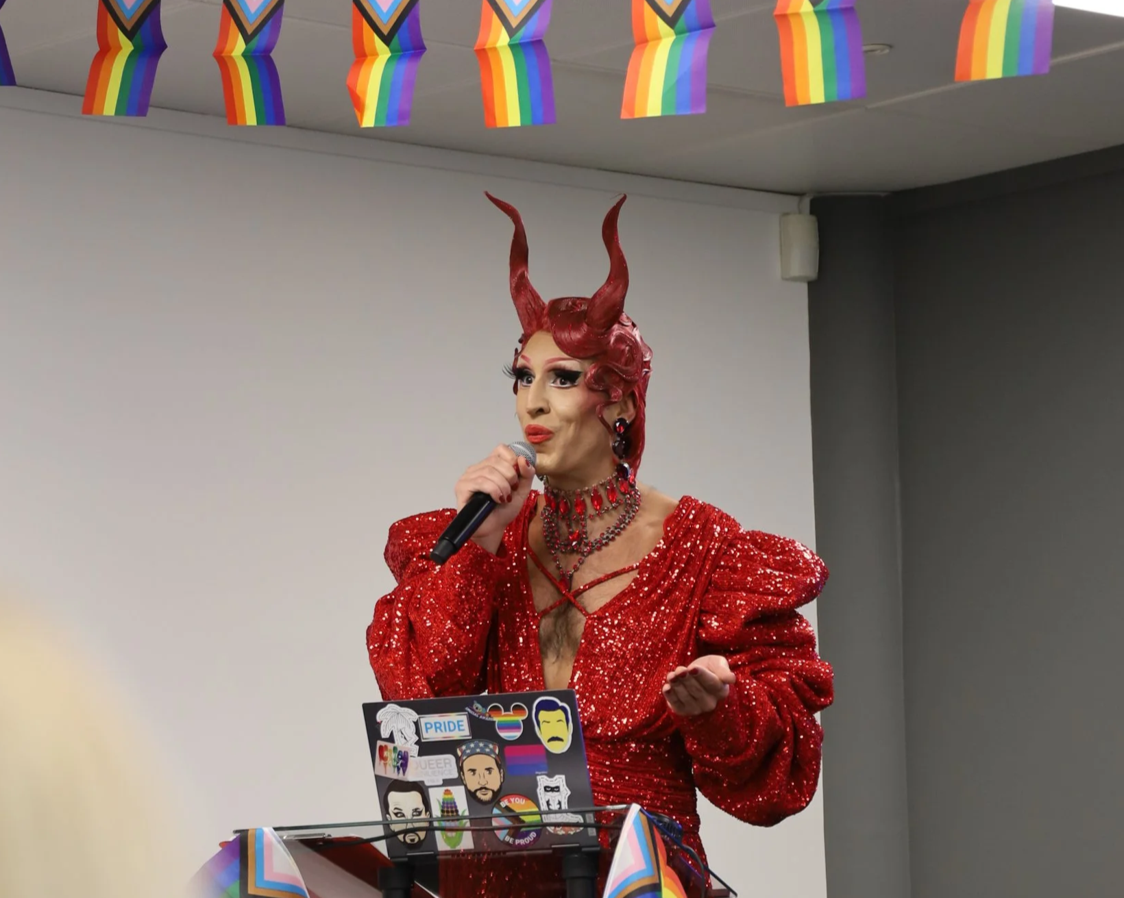 A person dressed as a devil with red horns, red lipstick, and dramatic makeup, wearing a sparkling red gown with puffed sleeves, speaking into a microphone at a Pride event, with rainbow flags hanging above and pride stickers on the laptop in front of them.