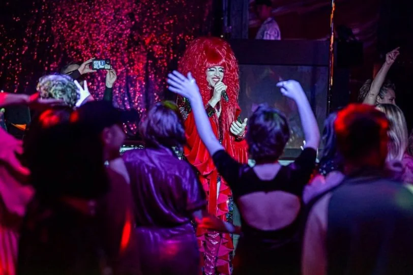 Drag performer in red costume singing on stage, surrounded by an enthusiastic audience with colorful lighting and a glittery backdrop.