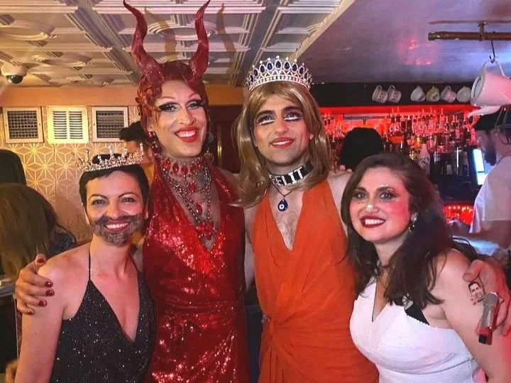 Four people in extravagant costumes at a party, including crowns, glittery outfits, and horns, posing and smiling in a bar with dim lighting.
