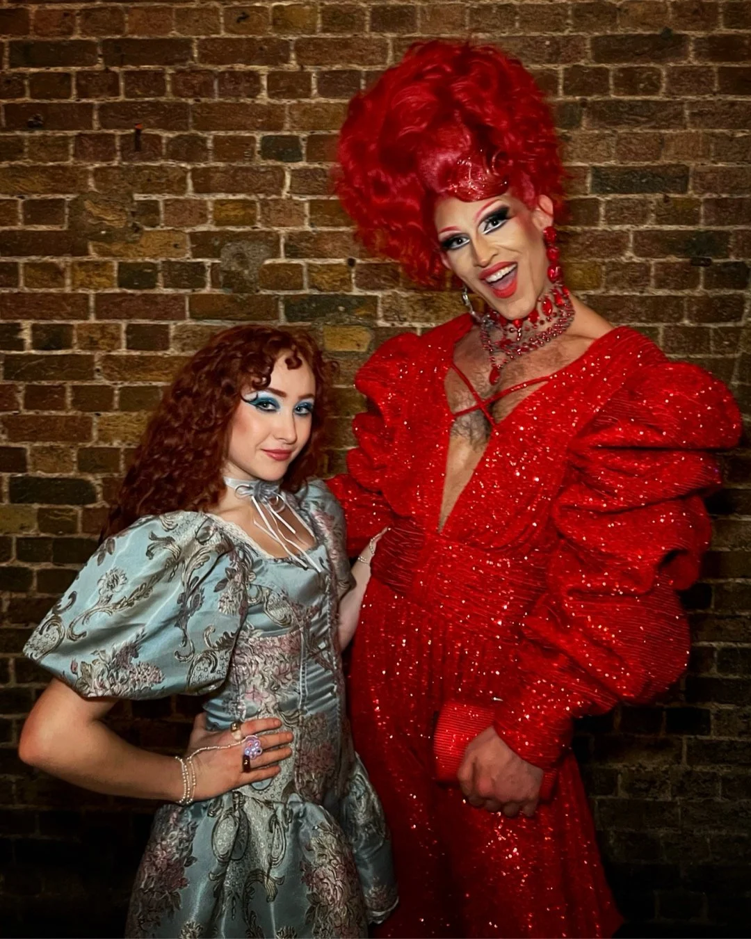 Two drag queens, Chappell Roan and Crayola the Queen, wearing colorful costumes against a brick wall. One in a floral patterned dress, the other in a red glittery outfit with a large hairstyle.