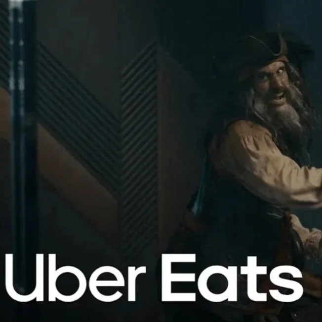 A man with long hair and a beard, dressed as a pirate, smiling in a dimly lit room with a wooden wall background. The text 'Uber Eats' is overlaid on the image.