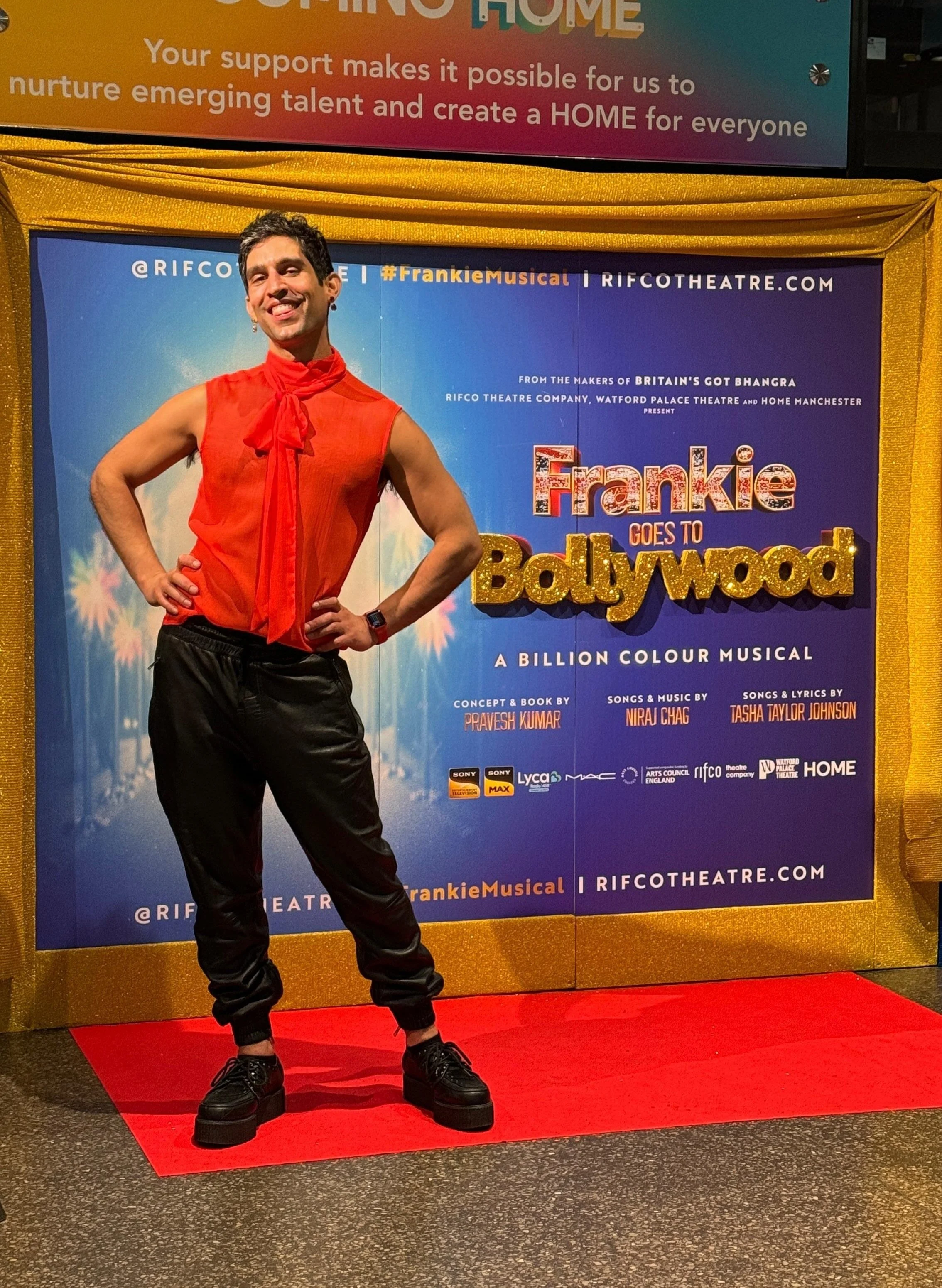 Person in a sleeveless orange shirt posing in front of a colorful poster for 'Frankie Goes to Bollywood' musical, standing on a red carpet.