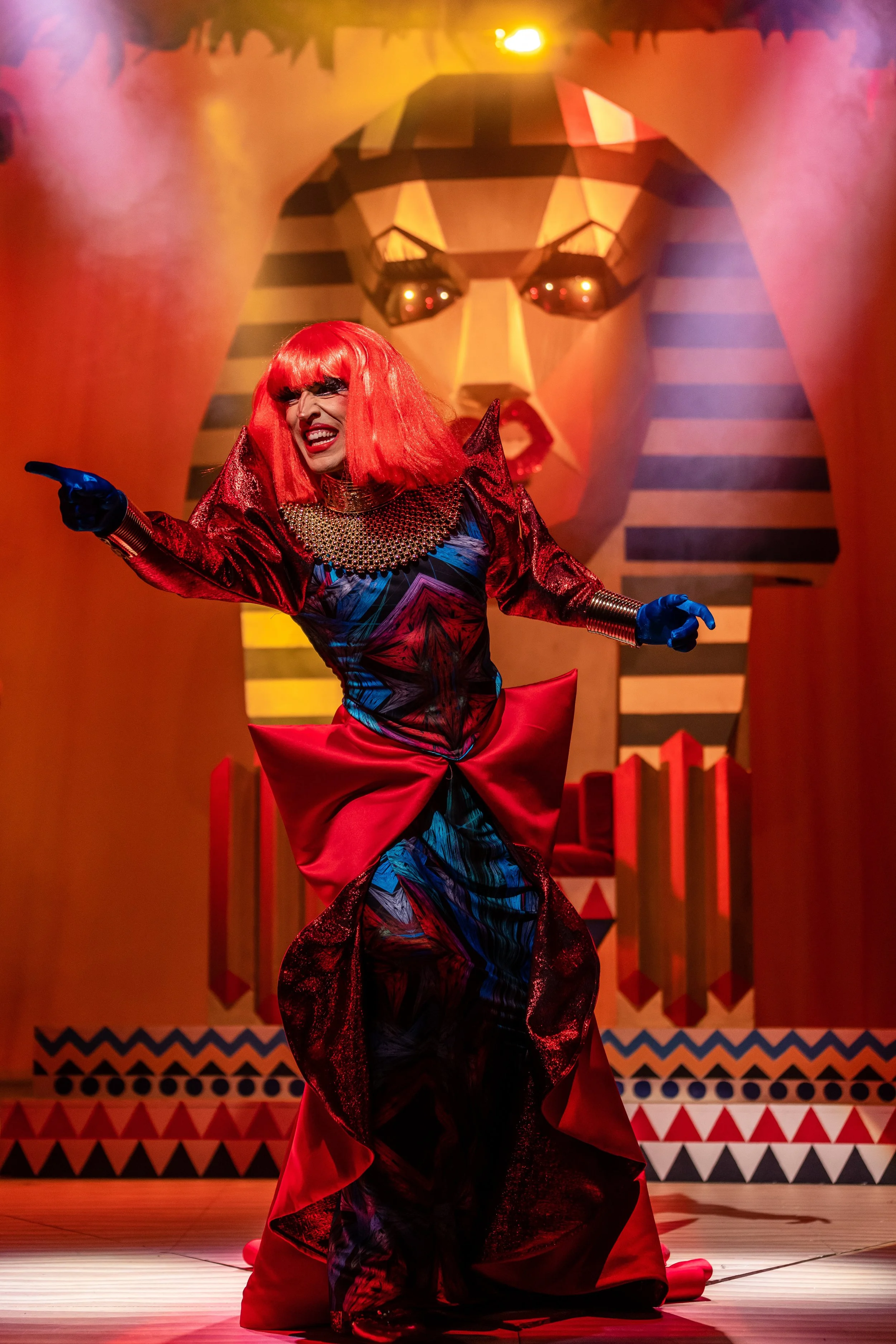 Performer in vibrant costume with red wig and blue gloves on stage with Egyptian-themed backdrop.