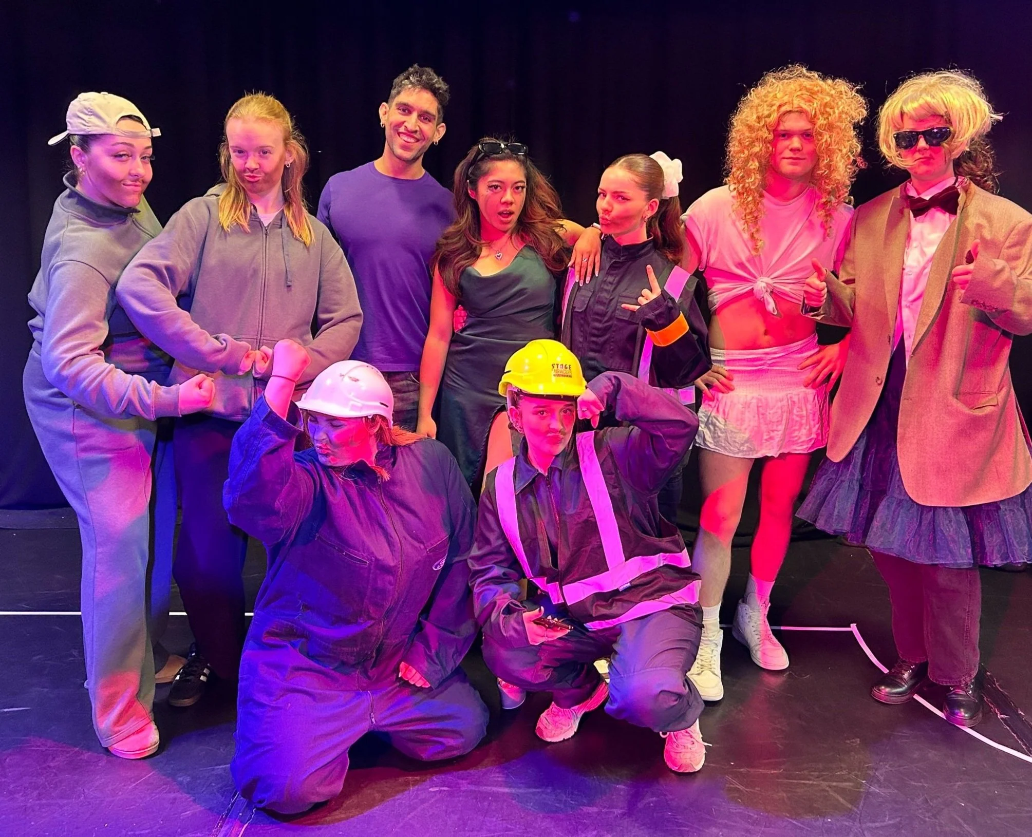 Group of people in theatrical costumes on stage, posing playfully, with various outfits including workwear and humorous attire.