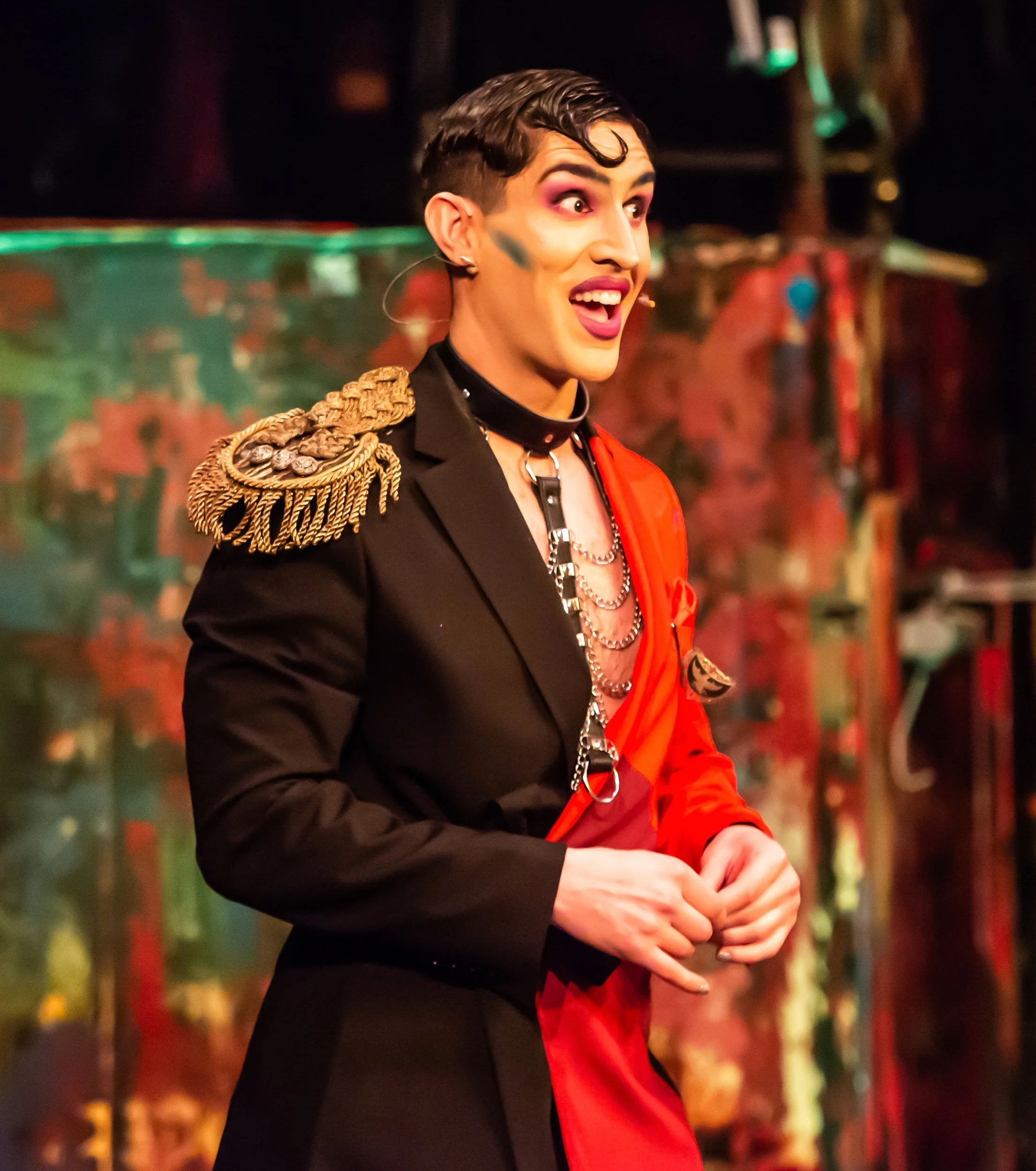 Person in colorful theatrical costume with makeup and accessories, standing on stage.