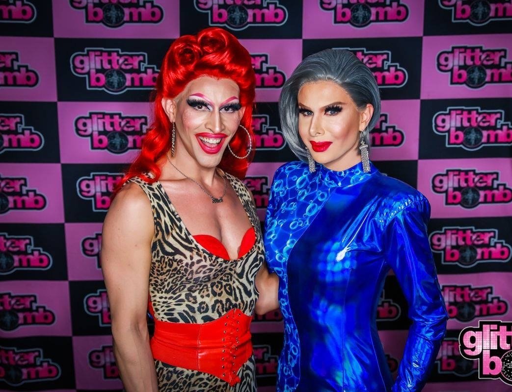 Crayola and Trinity the Tuck, in drag outfits posing in front of a "glitter bomb" backdrop. One is wearing a leopard print dress with red details and the other is in a metallic blue outfit. Both have bright makeup and styled hair.