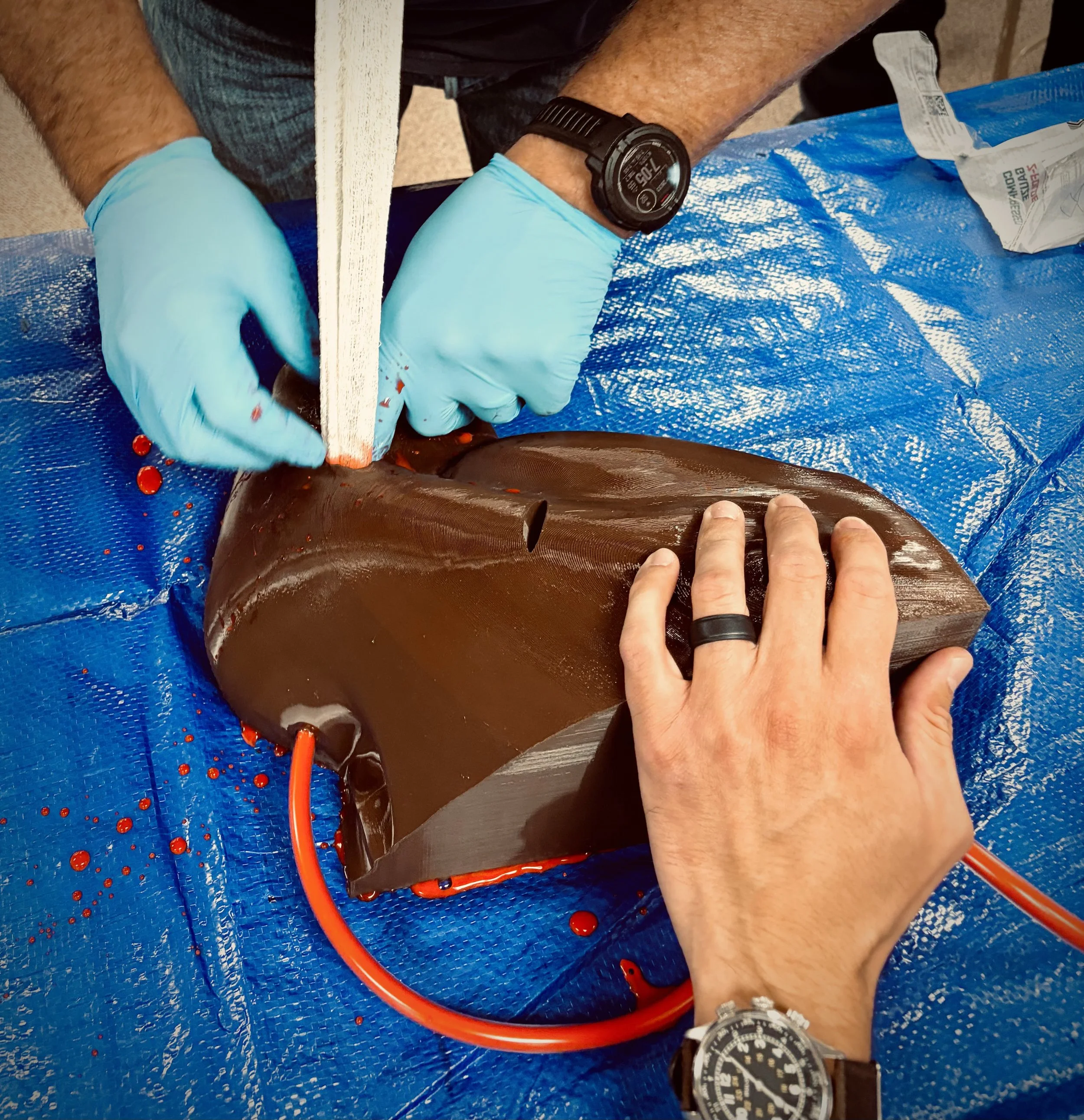 A person wearing blue gloves and a black watch is examining or working on a dark brown plastic or resin object on a blue tarp. There are red liquid drops and a red tubing nearby. The person is wearing a black shirt and silver wristwatch, and has a silver ring on the right hand.