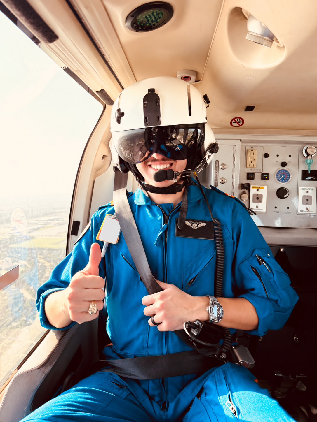 A smiling pilot in a blue aviation jumpsuit giving a thumbs-up inside the cockpit of an aircraft, wearing a white helmet with a visor and a headset.