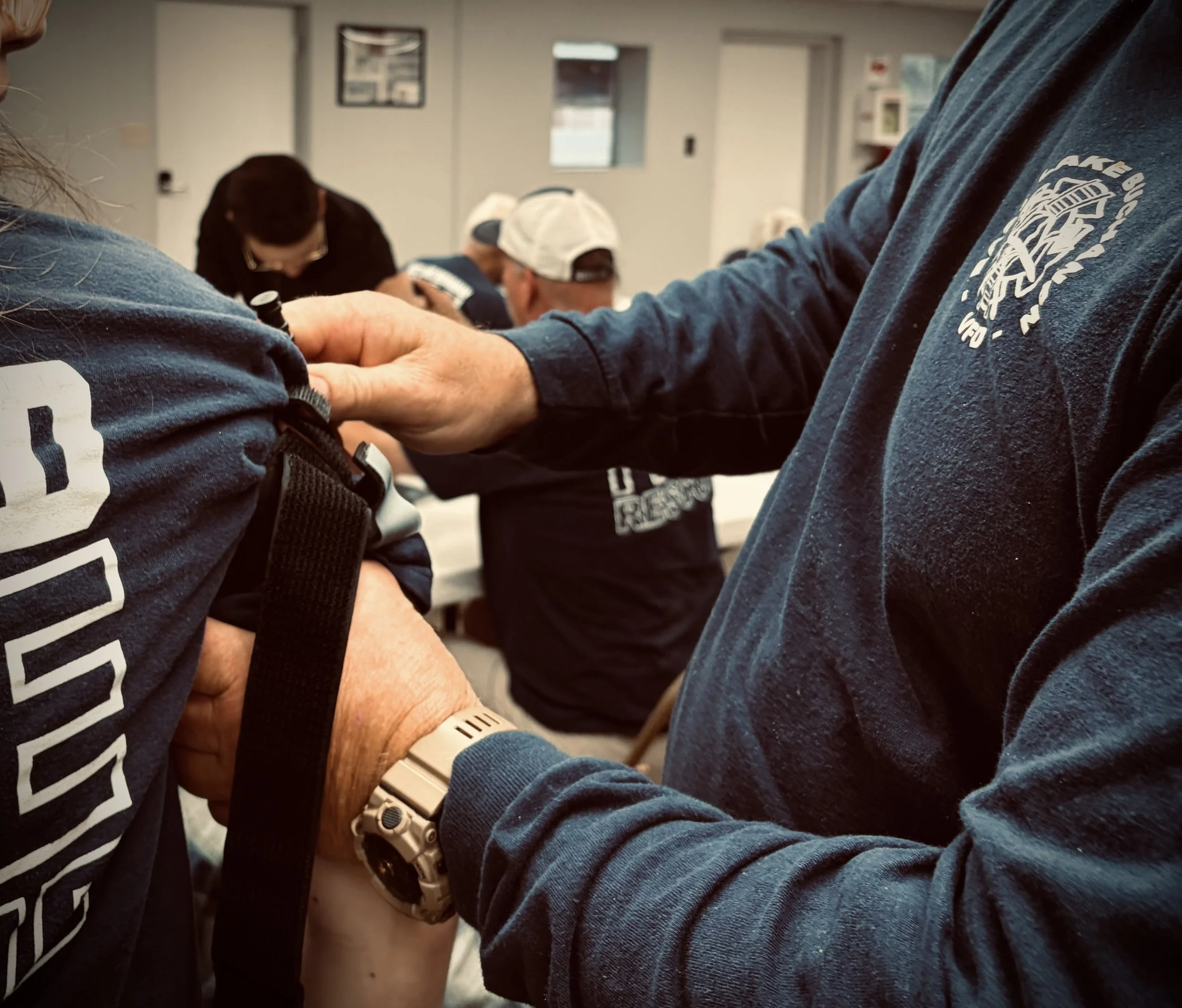 Close-up of two people holding hands, one wearing a blue long sleeve shirt with a patch that says 'Fire Rescue,' and the other wearing a black jacket with white geometric patterns, in a room with other people in the background.