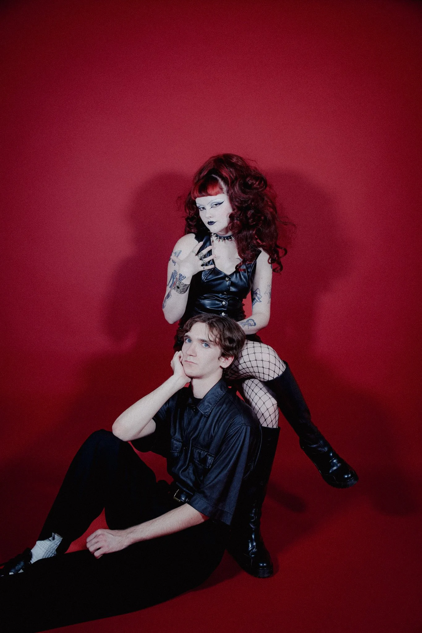 Two queer people sit in front of a red backdrop. One person is sitting in a chair while the other sits with their back against the other person on the ground. Both people are wearing all black with a goth aesthetic.