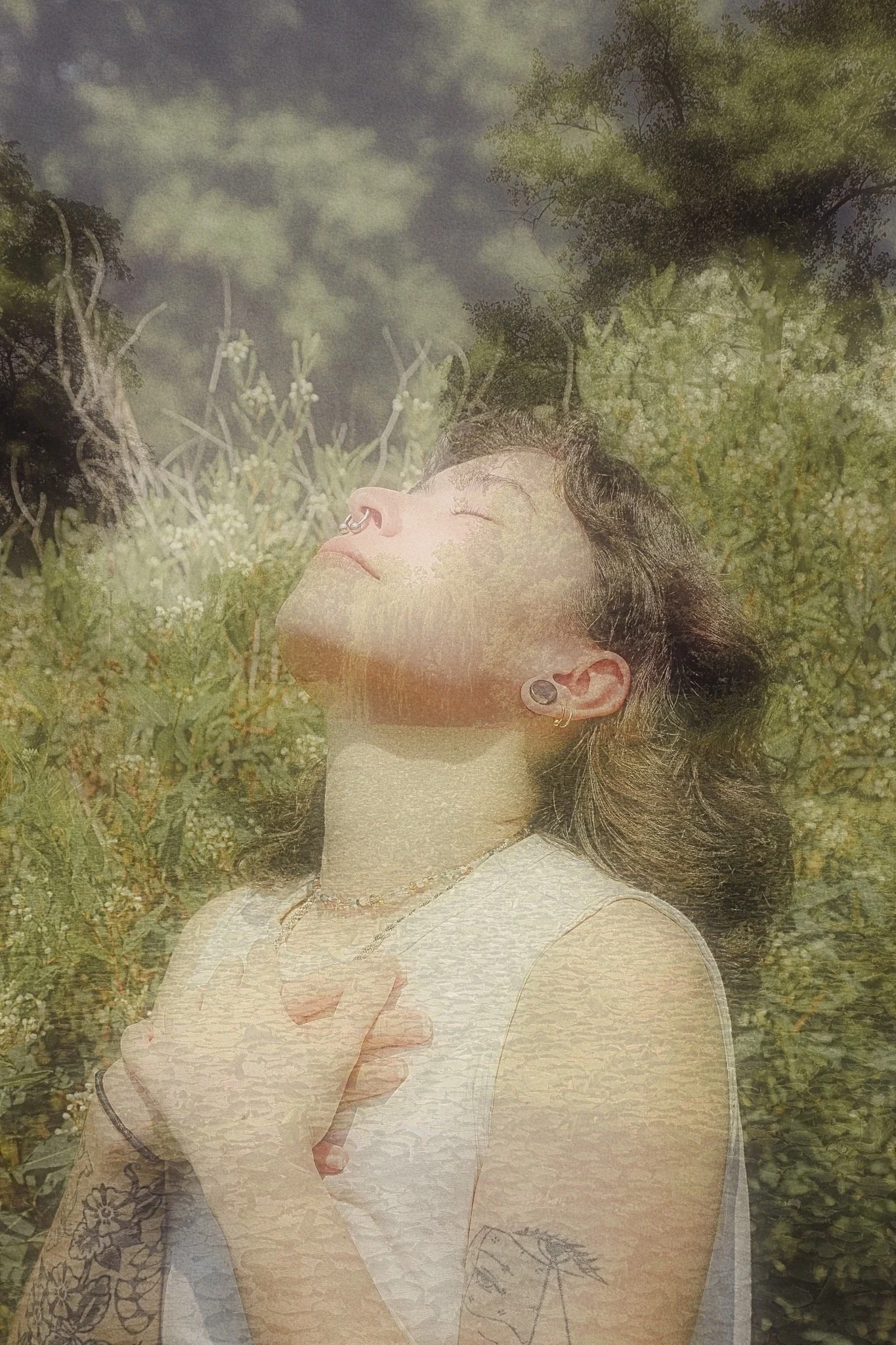 A queer person with a white top, brown curly hair and a nose piercing holds their hands close to their chest while looking up at the sky. They are surrounded by a green field and the photos had a slightly blurry effect on it.
