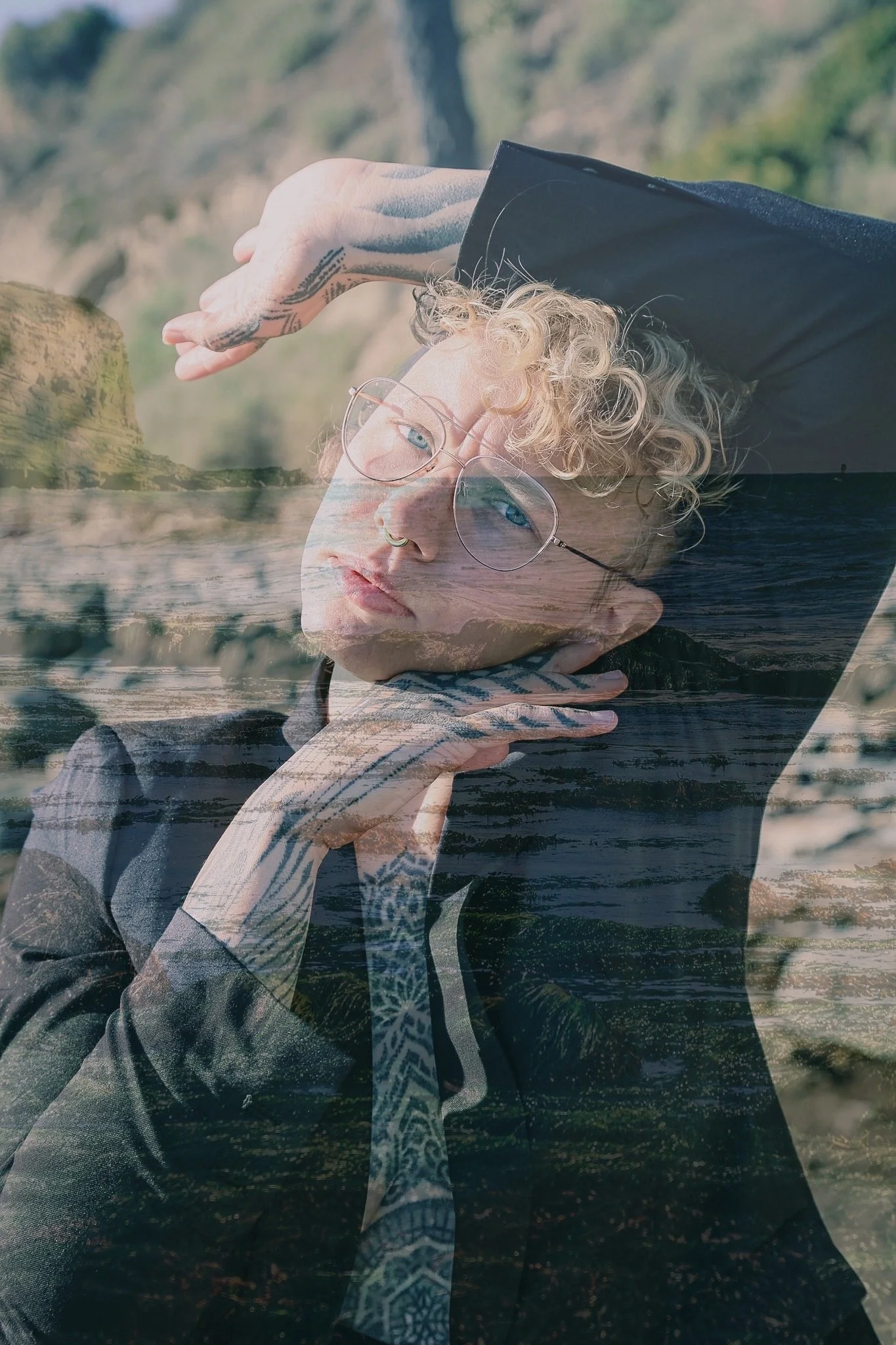 A lesbian portrait where one hand is on their face and one is over their head. They have curly hair and are wearing a black tux with tattooed hands and chest.