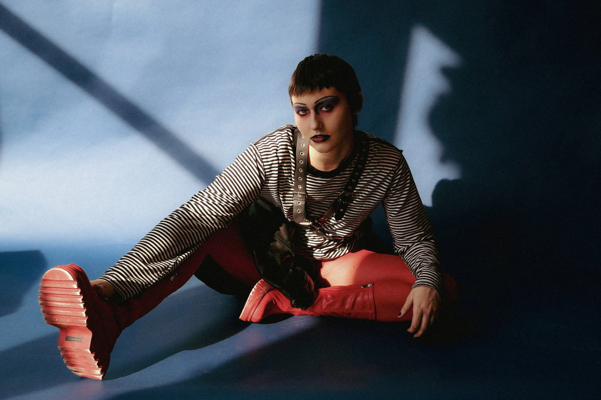 A queer person with blue, pink and black makeup sits seriously in front of a blue background. Wearing a leather jacket, striped black & white shirt, as well as red boots.