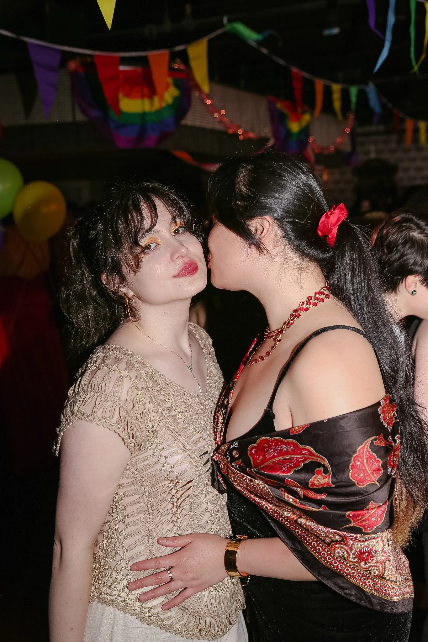 Two queer people stand in a bar in front of Pride themed decorations. One person looks directly at the camera and is wearing a mesh cream color top while the other kisses their cheek. The other person has black long hair and is wearing a black top.
