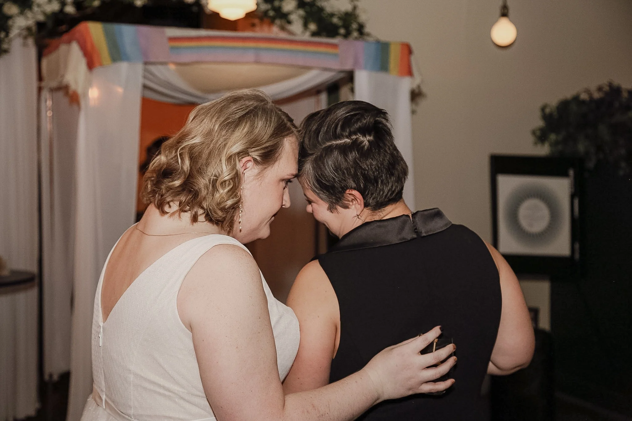 Two queer people who have just been married stand in front of a chuppah after their ceremony. One person is wearing a black and white pantsuit while the other wears a white dress. One has their head resting on another.