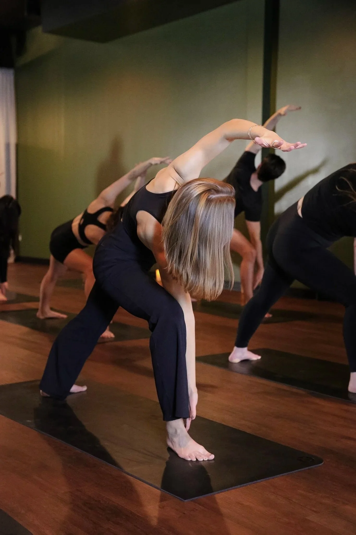 A person bends with their hand over their head on their yoga mat within a yoga studio. The person has on a black activewear set and blonde hair. The space has green walls and brown wooden floors. 