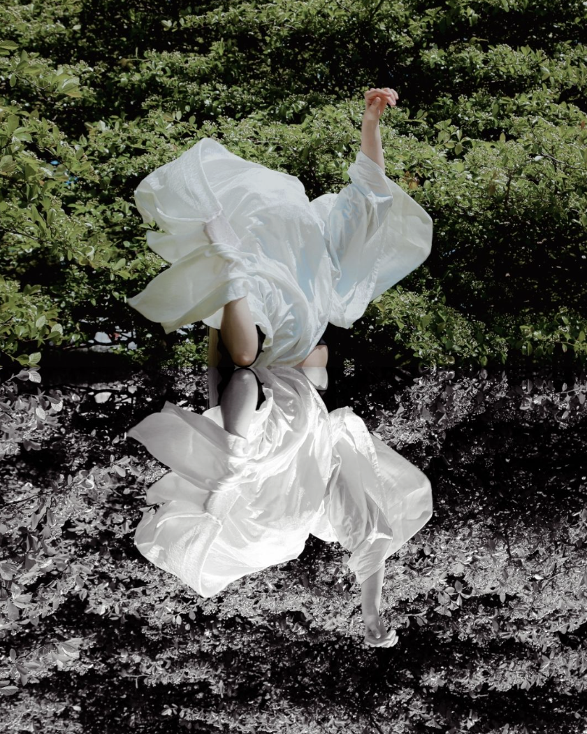 A person has their hands up in front of a tree with a sheet flowing over their face