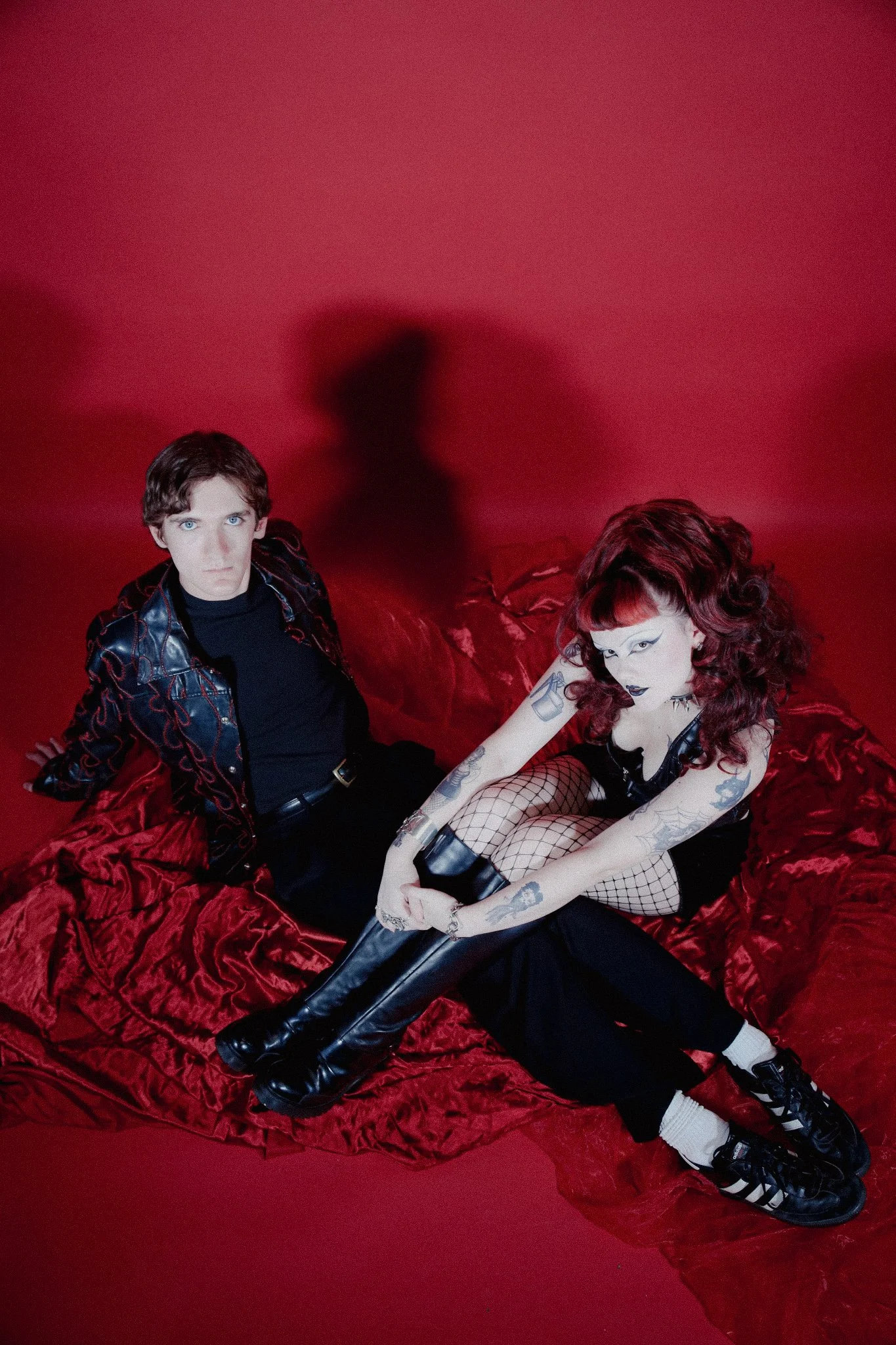 Two queer people sit side by side in front of a red background. They are sitting on a red silk blanket with serious expressions. Both people are wearing all black with a goth aesthetic.