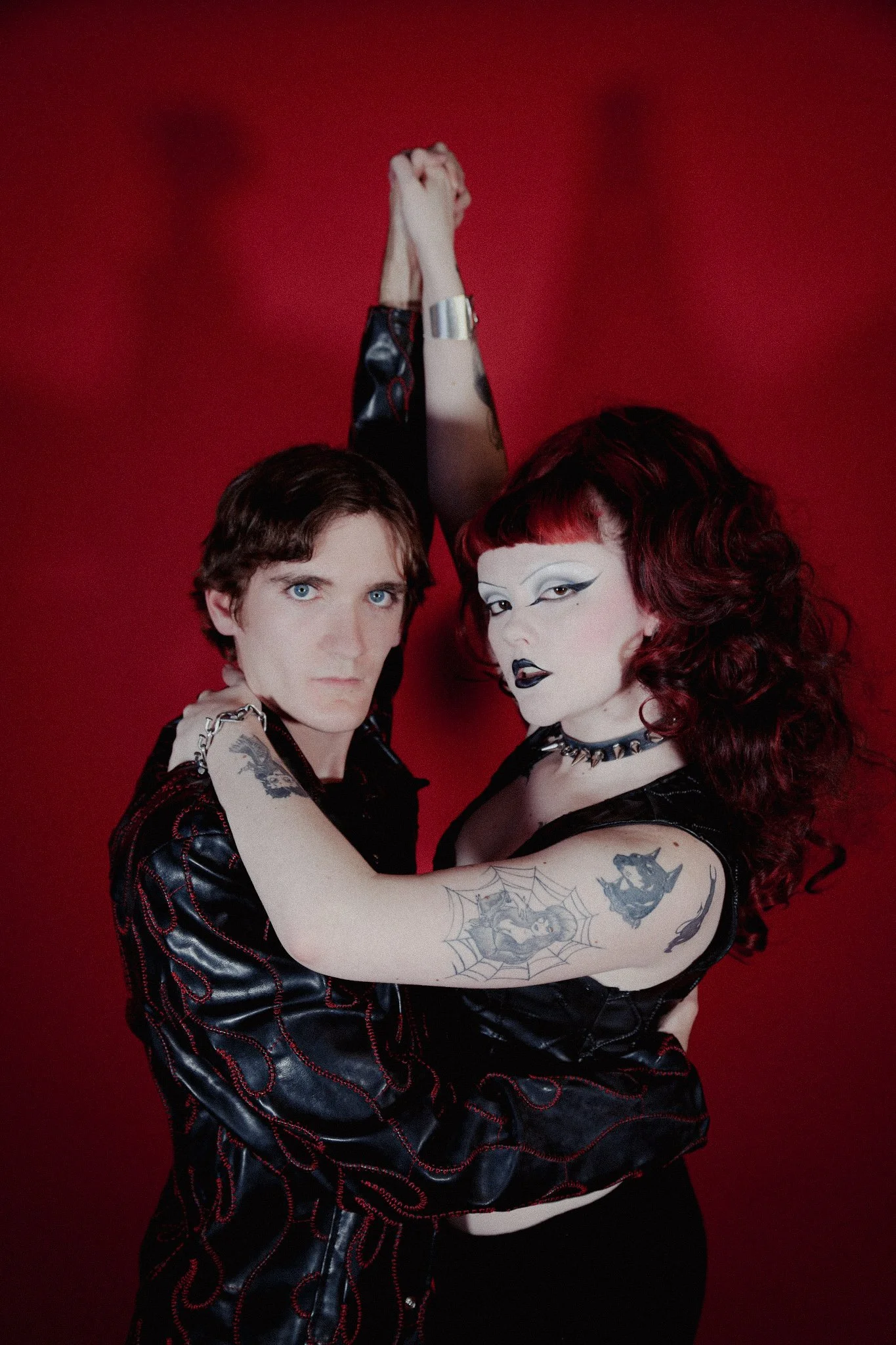 Two queer people wearing all black with a goth-like aesthetic stand in front of a red backdrop holding hands above their heads. They both have a serious facial expression.