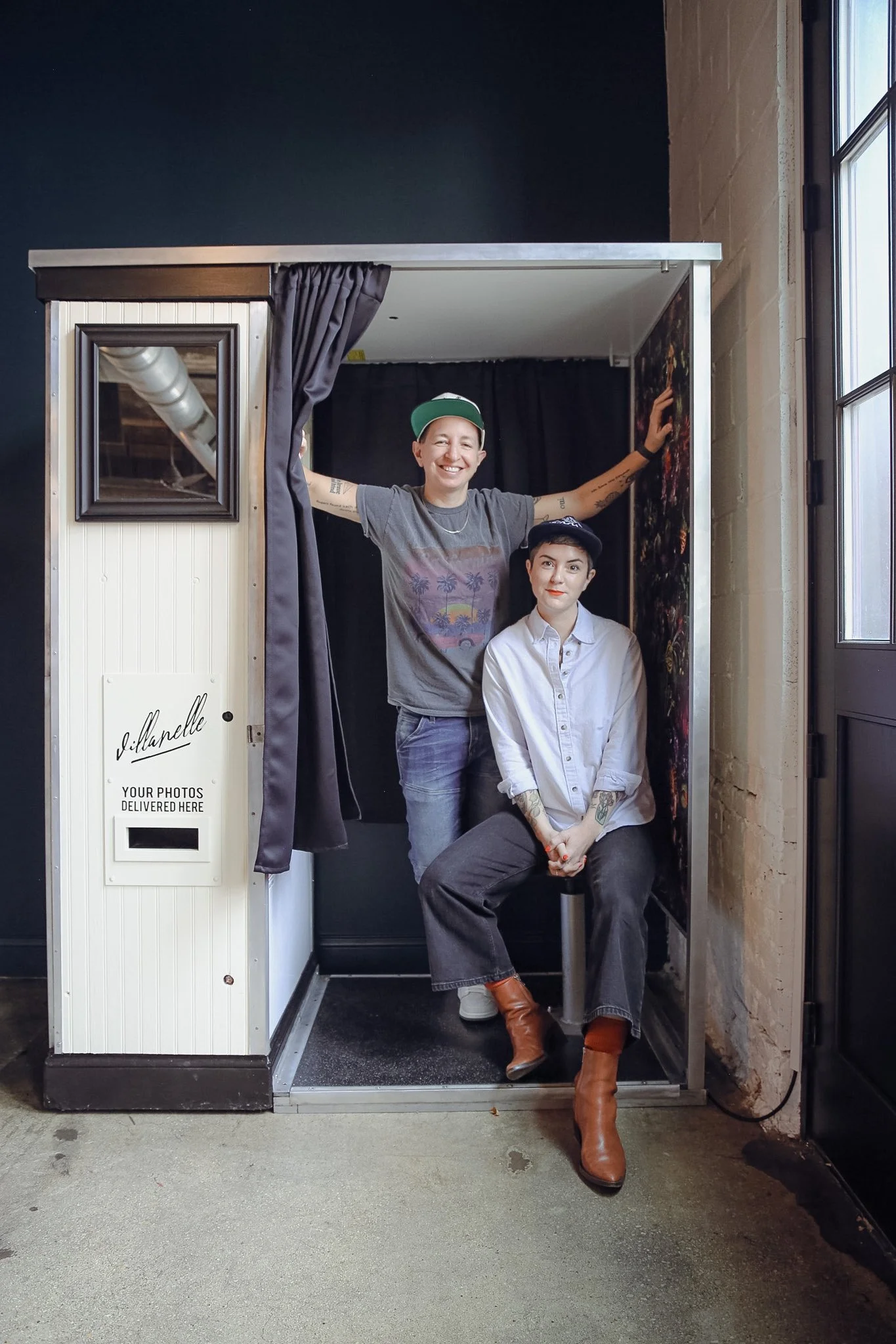 A photo contains two queer people sitting in a photo booth smiling. The label on the side of the photo booth says "Villanelle". They are both wearing casual clothing. 