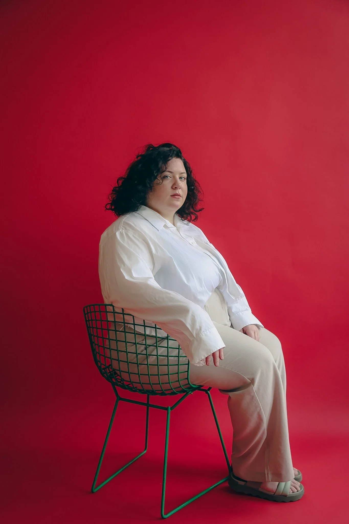 A queer person with black curly hair, white top and slacks sits on a green wire chair in front of a red background. They have a serious expression on their face and their hand is resting atop of the chair.